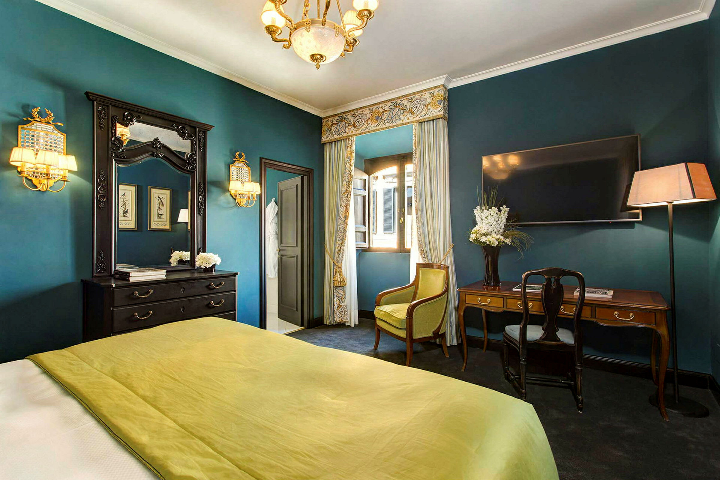 Et elegant innredet hotellrom med grønt sengeteppe, skrivebord, speil og gardiner foran et åpent vindu. (AI-generert tekst)