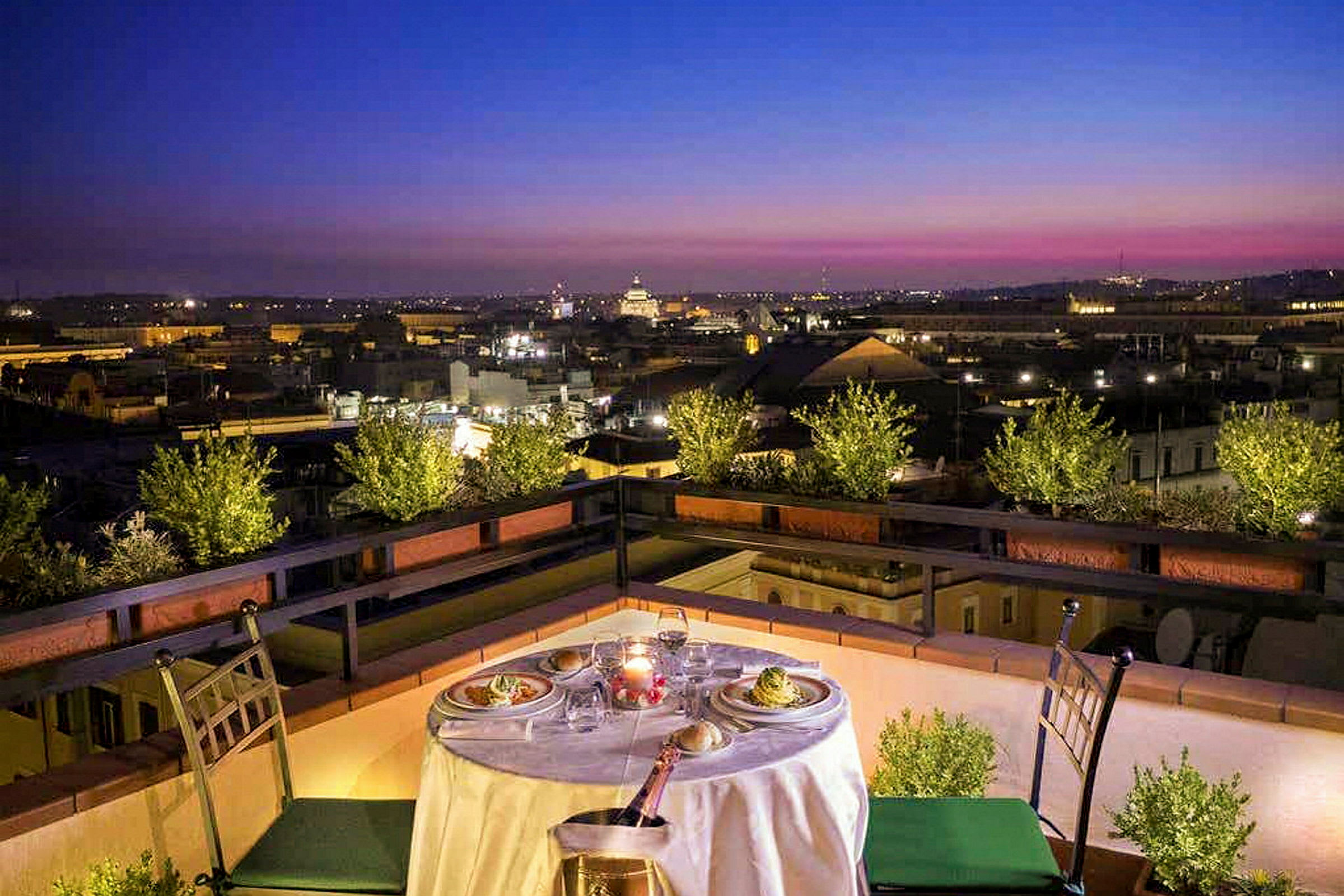 Et dekket bord med mat og drikke på en terrasse med utsikt over en by ved solnedgang. (AI-generert tekst)