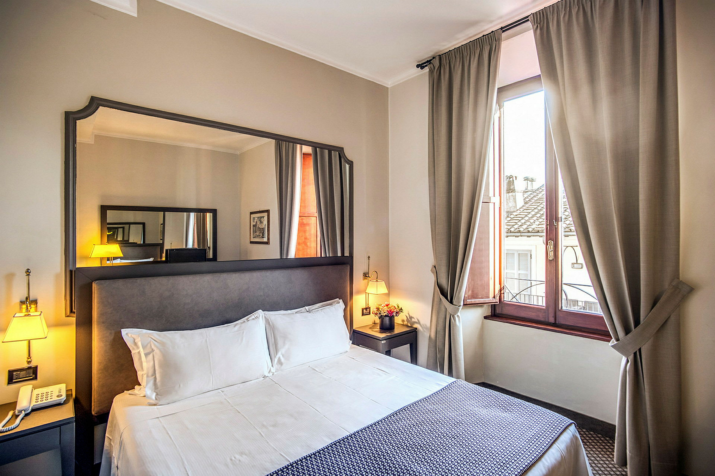 Hotellrom med dobbeltseng, nattbord, speil og vindu med beige gardiner. (AI-generert tekst)