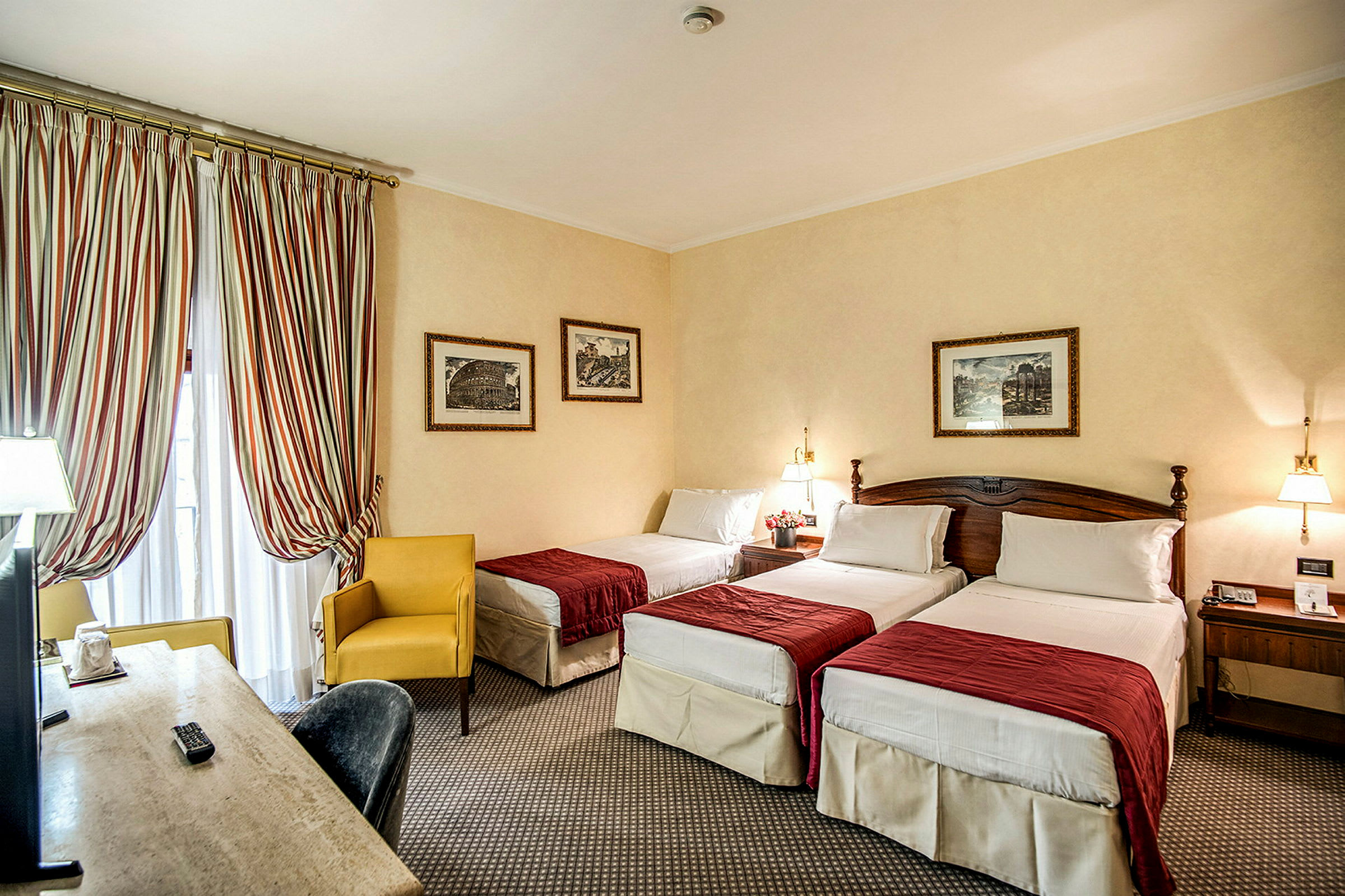 Hotellrom med tre enkeltsenger og rødt sengetøy. En gul lenestol ved vinduet samt et skrivebord med TV. (AI-generert tekst)