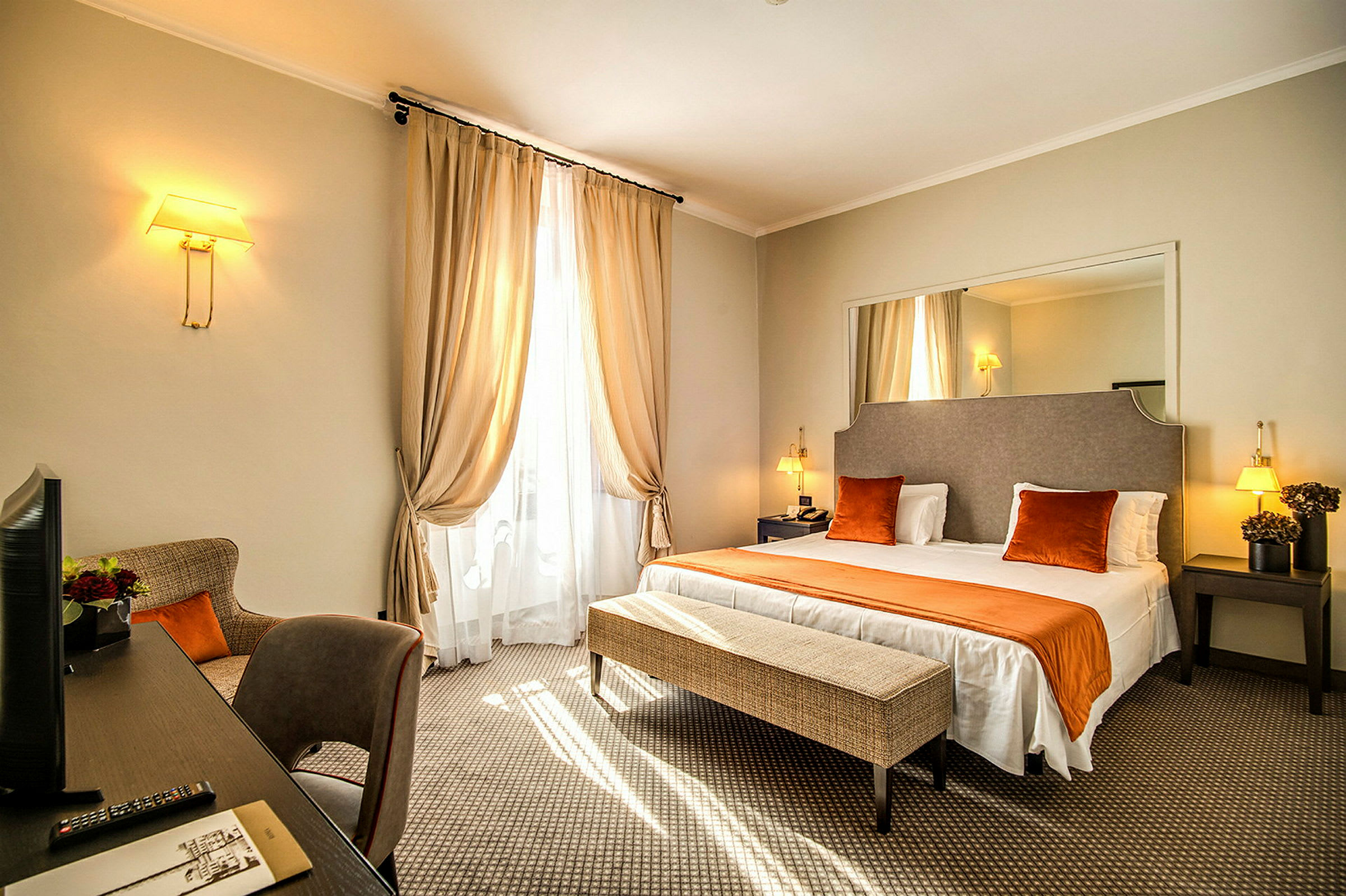 Et hotellrom med en dobbeltseng, nattbord og en benk ved foten av sengen. Gardiner dekker vinduet, og det er et skrivebord med en stol ved siden av. (AI-generert tekst)