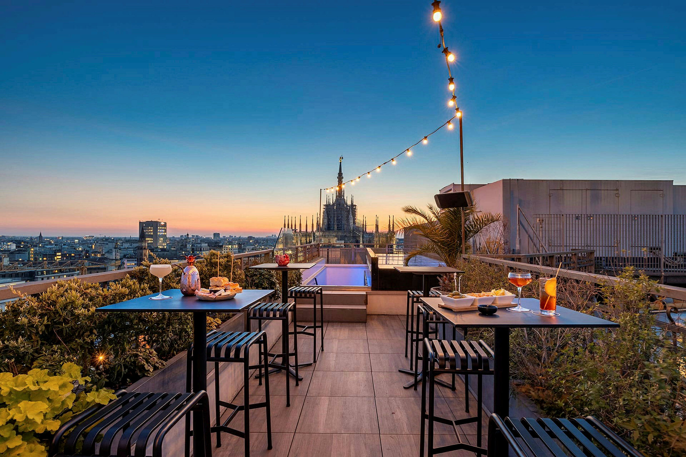 En takterrasse med utsikt over en by ved solnedgang, hvor små bord og stoler er plassert med drikke og snacks. (AI-generert tekst)