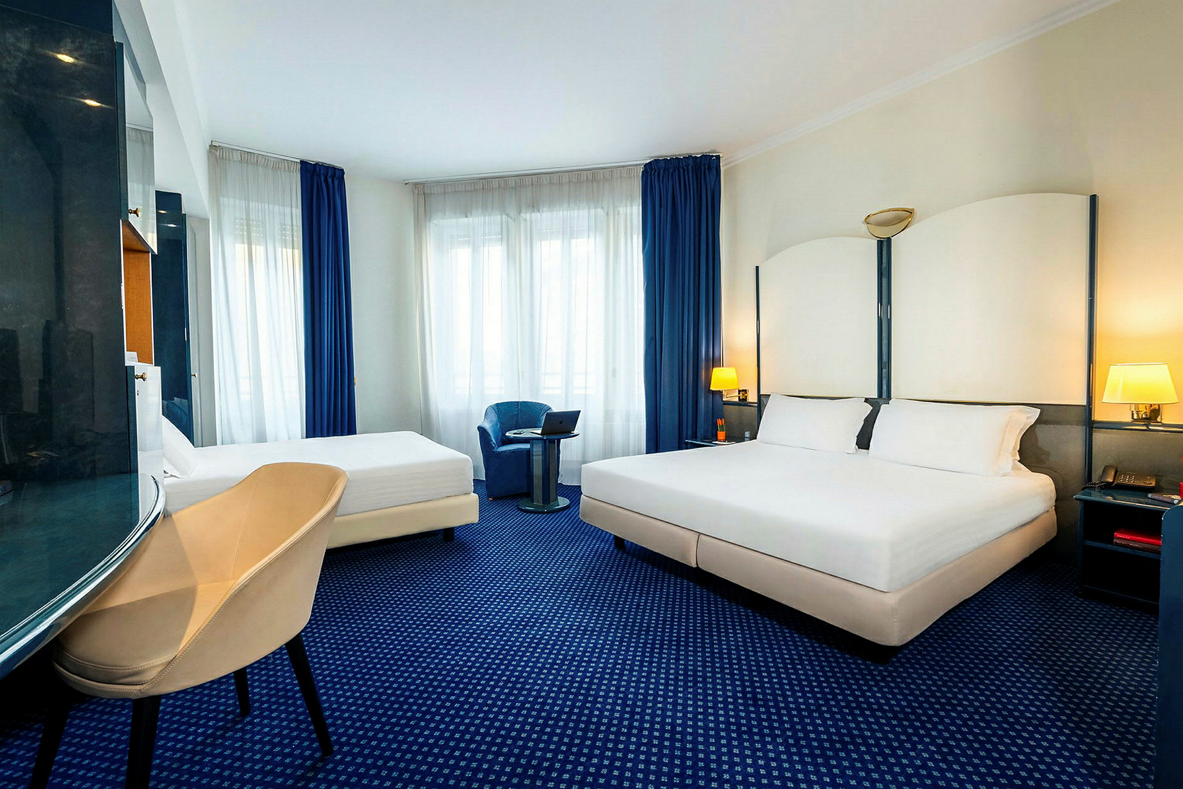 Hotellrom med en dobbeltseng og en enkeltseng, lenestol og vindu med blå gardiner. (AI-generert tekst)