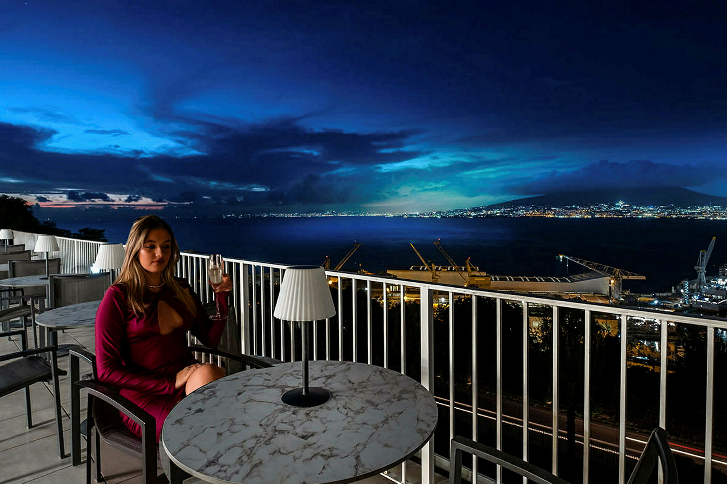 Kvinne sitter ved et bord på en terrasse med utsikt over et rolig havmiljø om natten. (AI-generert tekst)