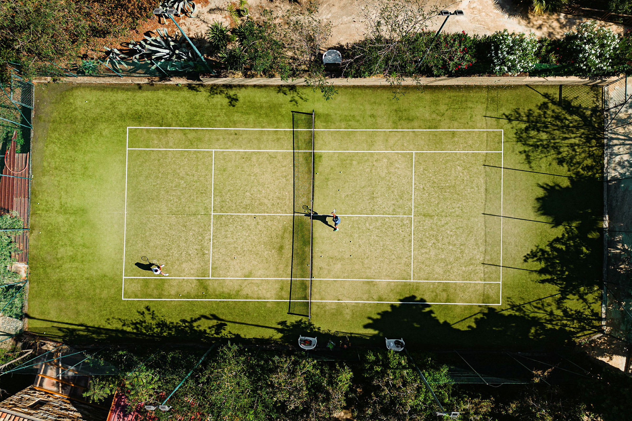 Luftfoto av to personer som spiller tennis på en grusbane omgitt av vegetasjon. (AI-generert tekst)