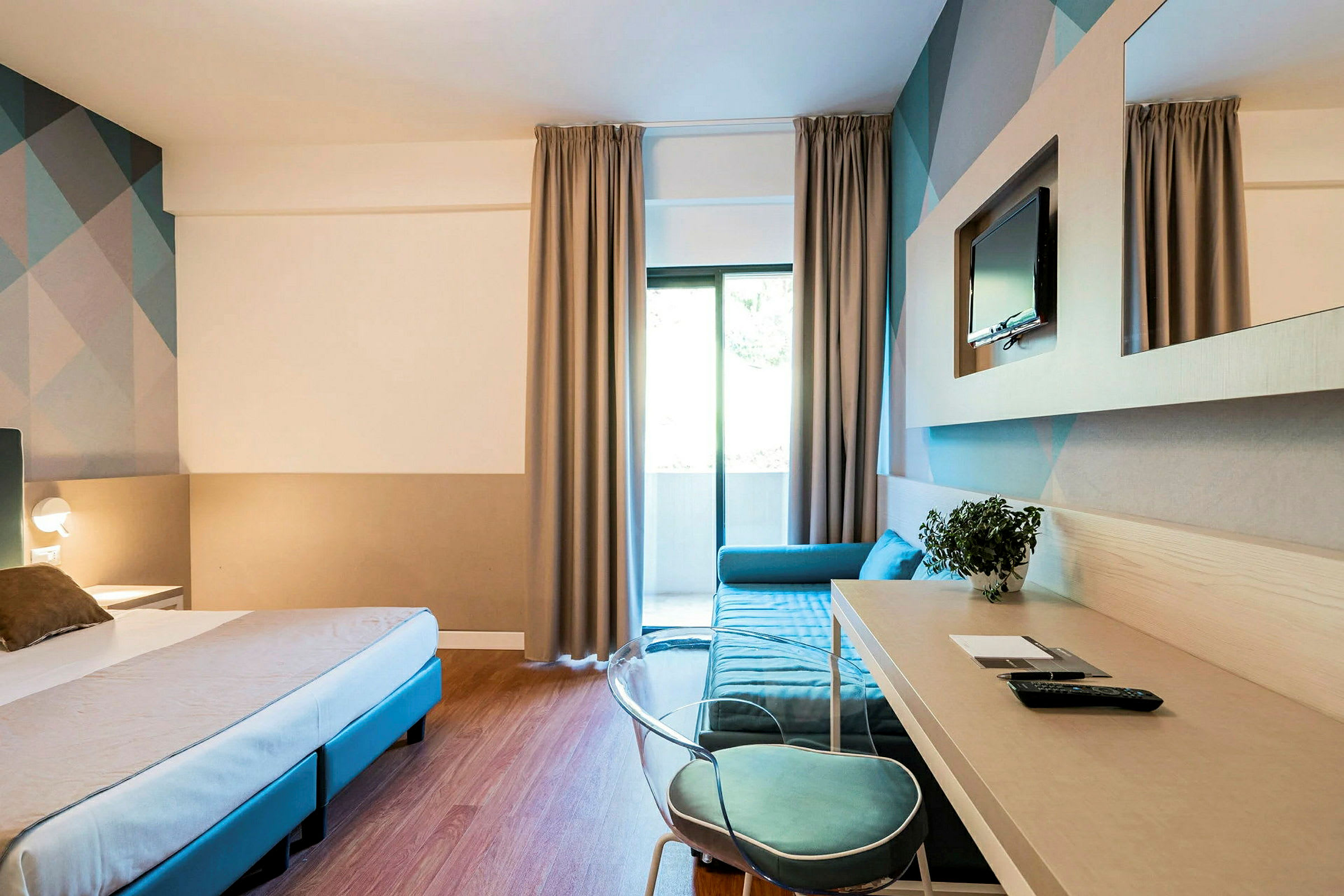 Hotellrom med seng, skrivebord og sofa, samt TV på veggen. (AI-generert tekst)