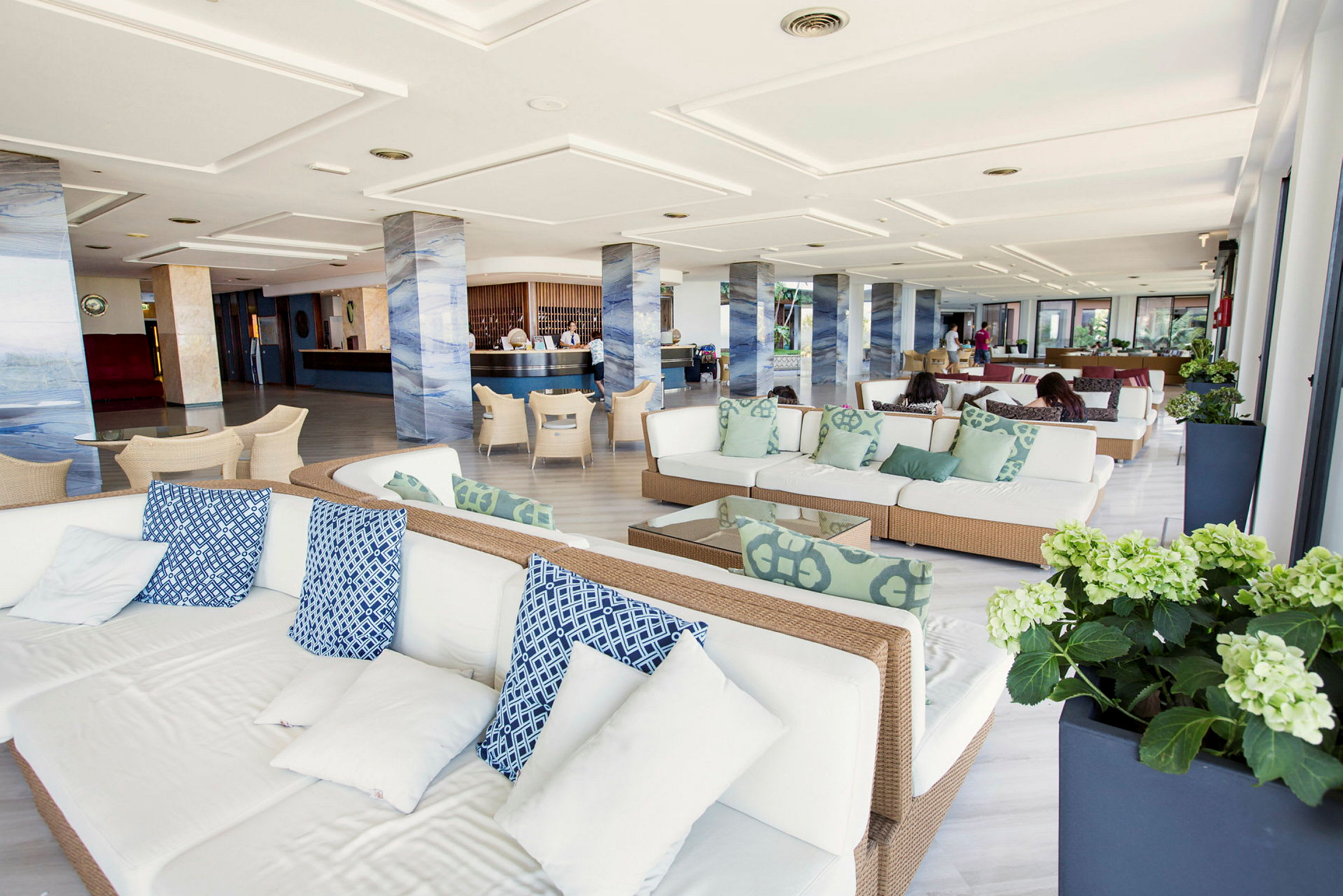 Et moderne lobbyområde med hvite sofaer og blå samt grønne puter. Området har søyler i en marmorert design og et resepsjonsområde i bakgrunnen. (AI-generert tekst)