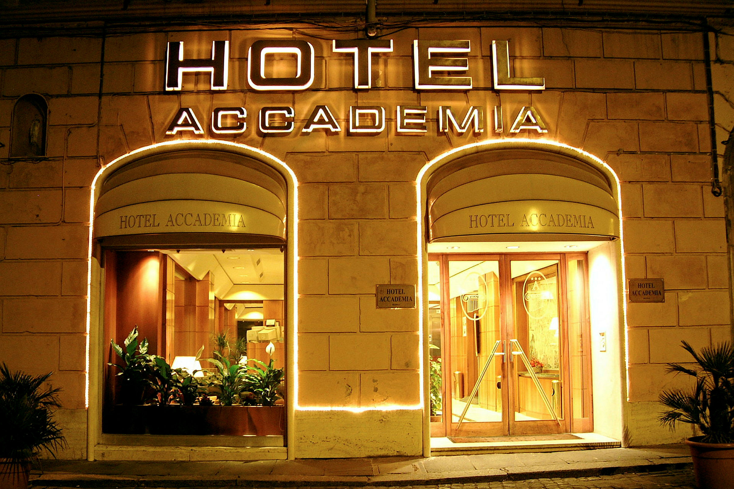 Hotel Accademias opplyste inngang med glassdører og planter. (AI-generert tekst)