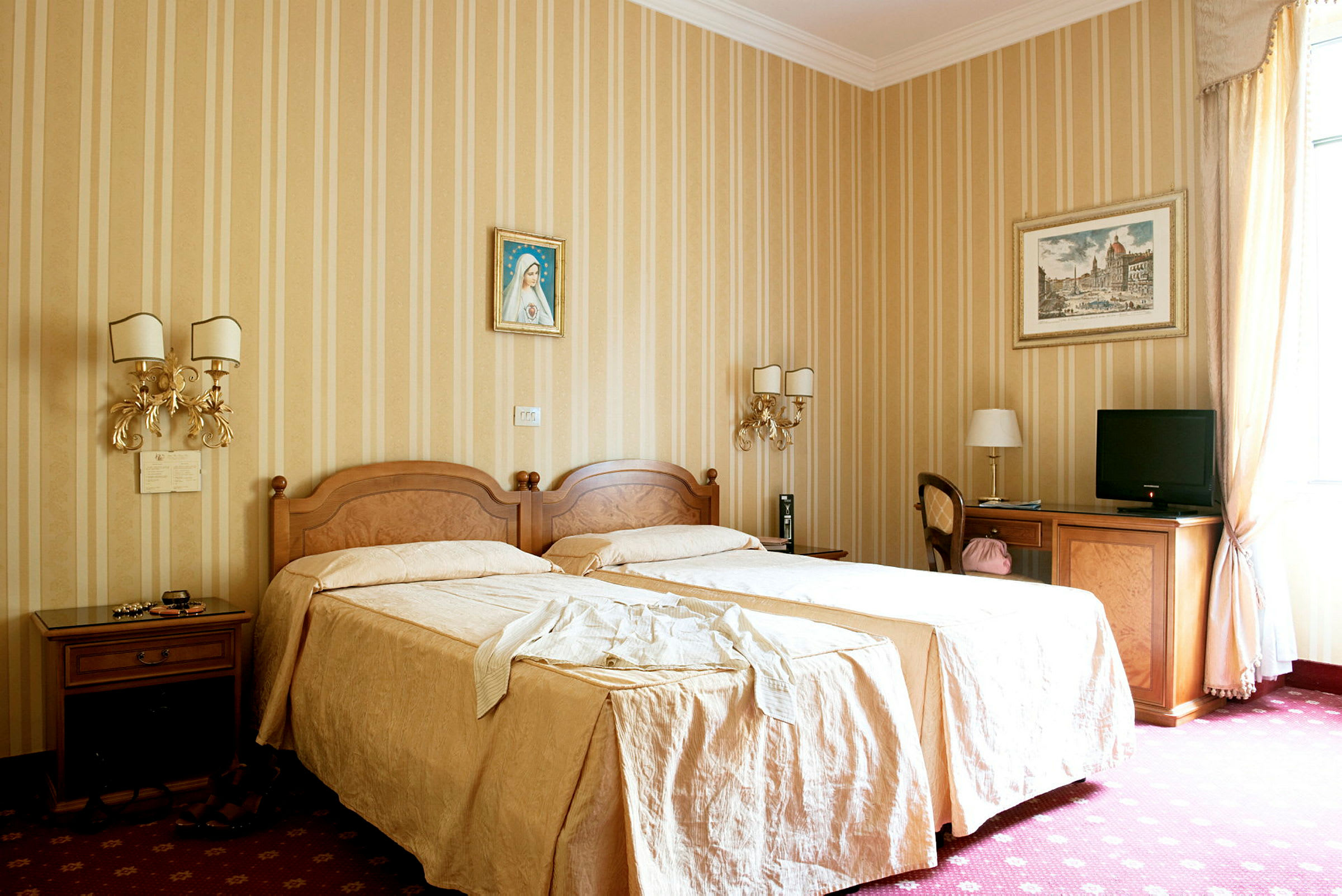 Et hotellrom med to enkeltsenger dekket med beige sengetøy, dekorerte vegger med malerier, og møbler inkludert bord, stoler og en TV. (AI-generert tekst)