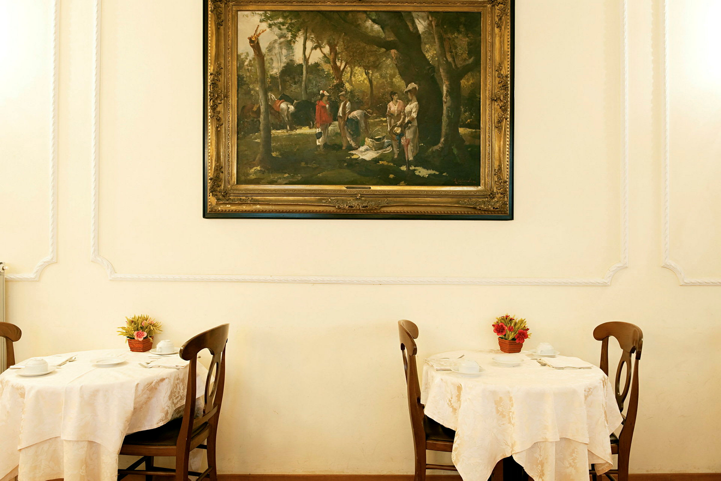 Et rom med to dekkede bord med stoler, dekorert med blomsterpotter og et stort innrammet kunstverk på veggen. (AI-generert tekst)