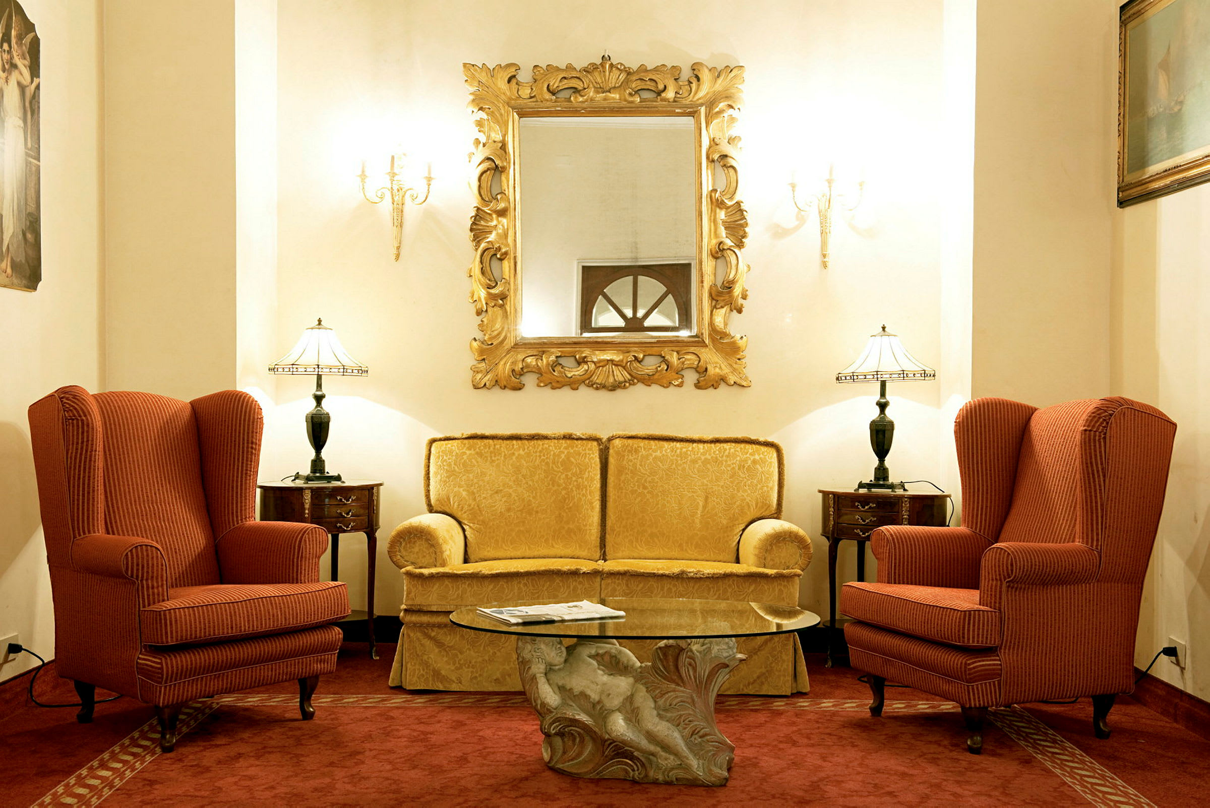 En hotellounge med to lenestoler, en sofa, et glassbord og et dekorert speil. (AI-generert tekst)