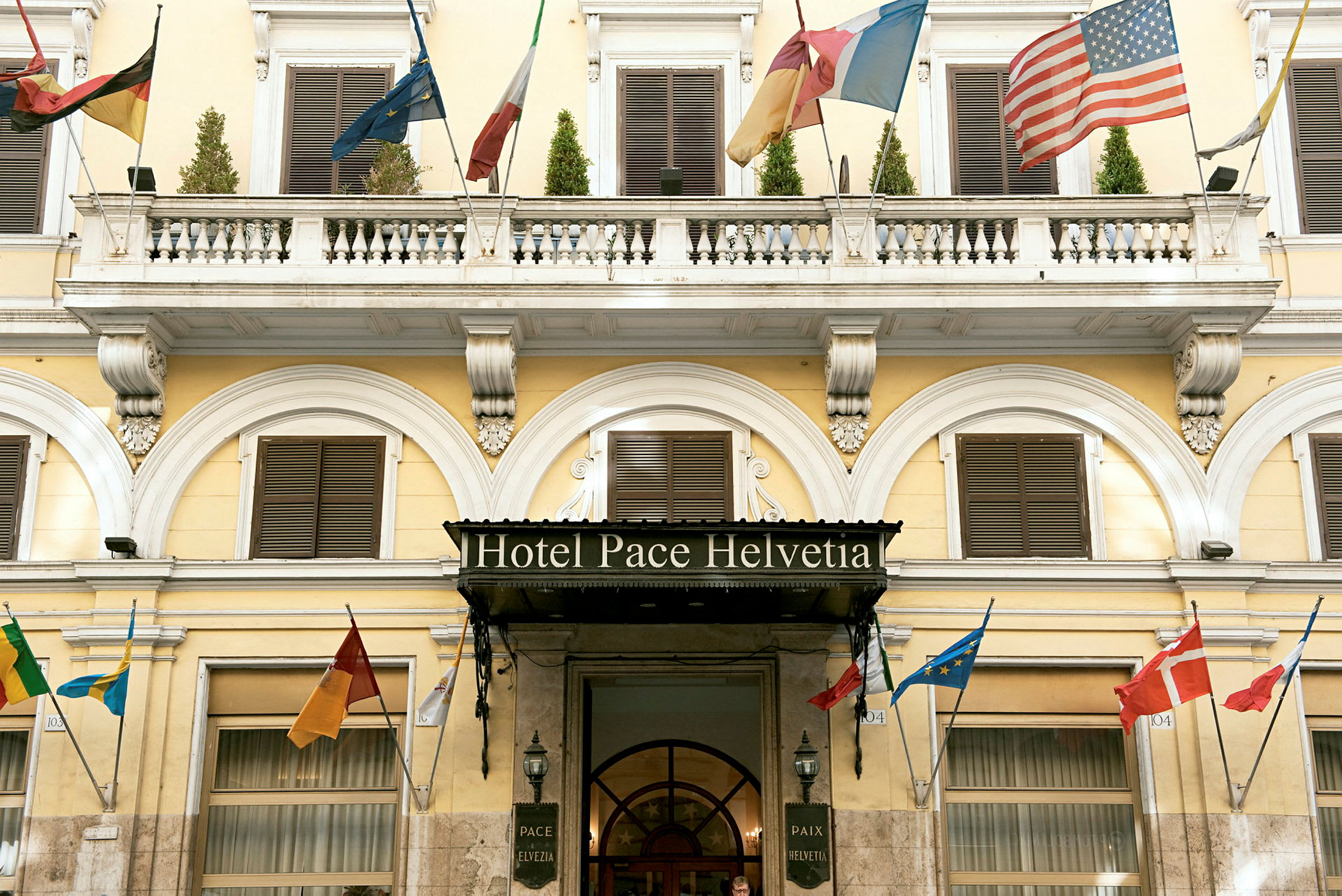 Hotellfasade med navnet 'Hotel Pace Helvetia', dekorert med flere lands flagg. (AI-generert tekst)
