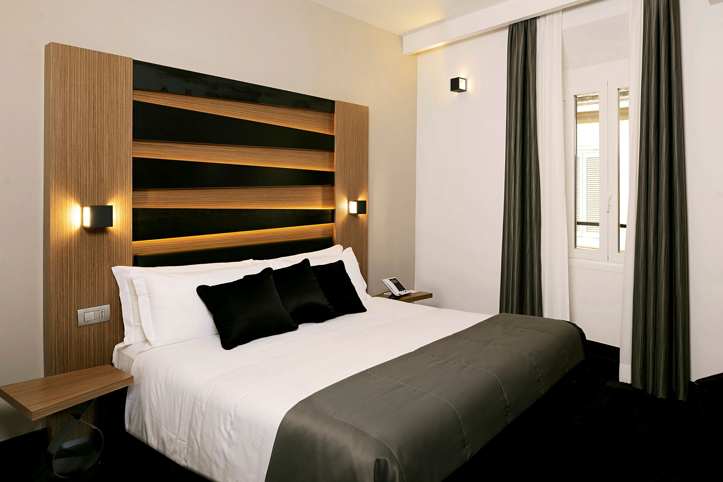 Dette bildet viser et hotellrom med en moderne og elegant seng med hvitt sengetøy og mørke dekorative puter. Rommet har en enkel og stilren design med et vindu med nøytrale gardiner. (AI-generert tekst)