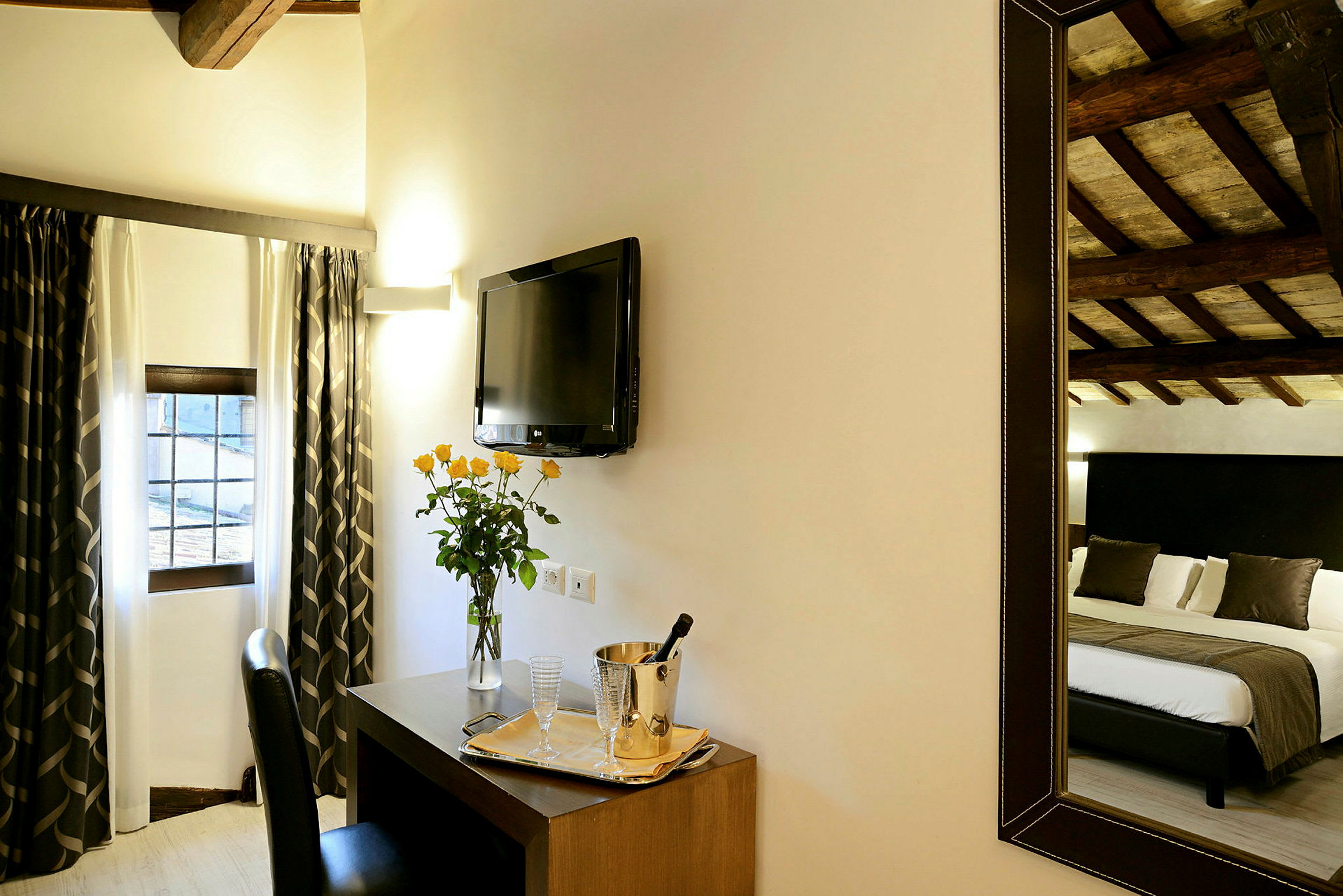 Et hotellrom med gardiner ved vinduet, en TV på veggen, et skrivebord med en vase blomster og serveringsbrett samt et speil som reflekterer en seng. (AI-generert tekst)