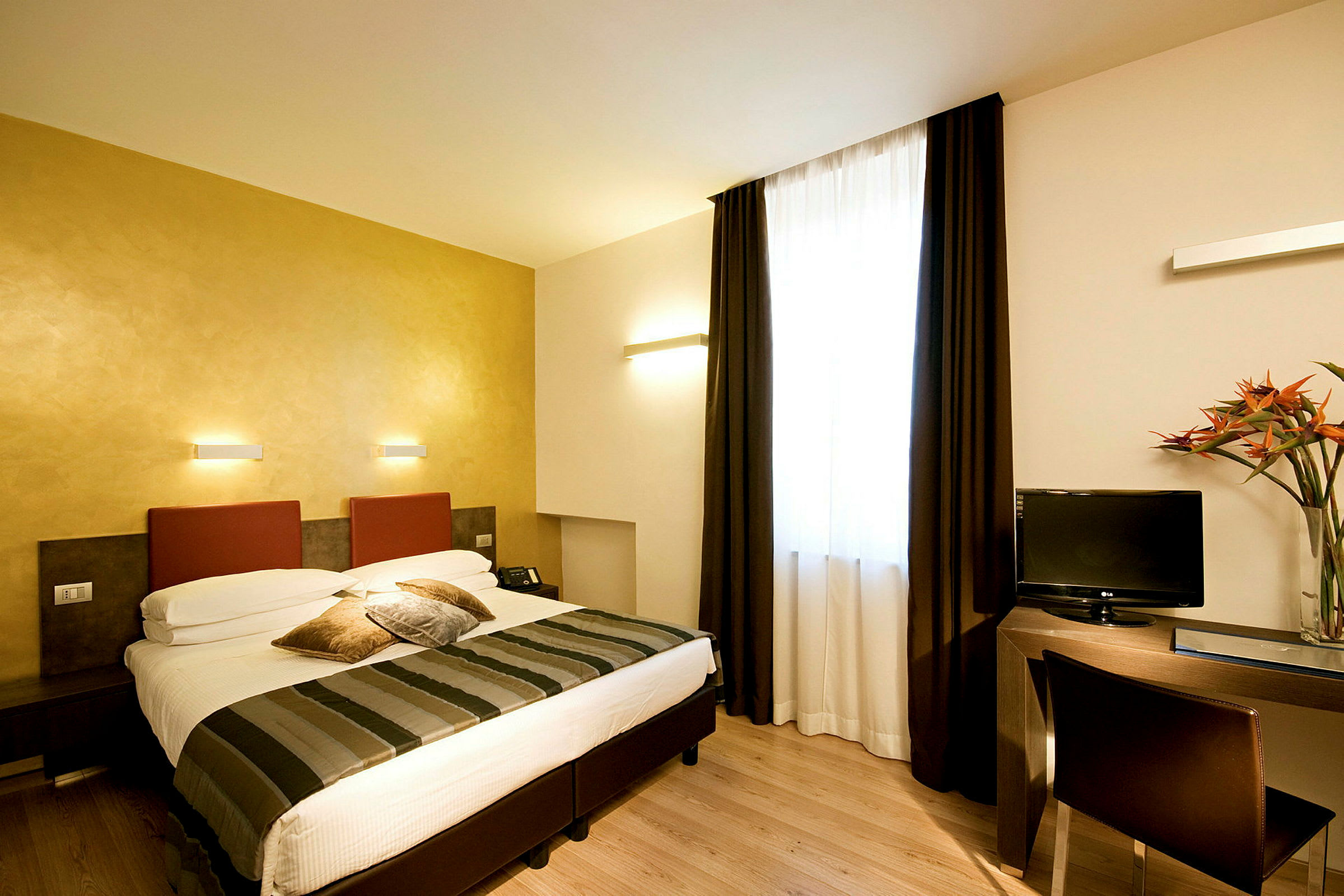 Et hotellrom med en dobbeltseng, skrivebord og TV. Rommet har lyse vegger og et stort vindu med mørke gardiner. (AI-generert tekst)