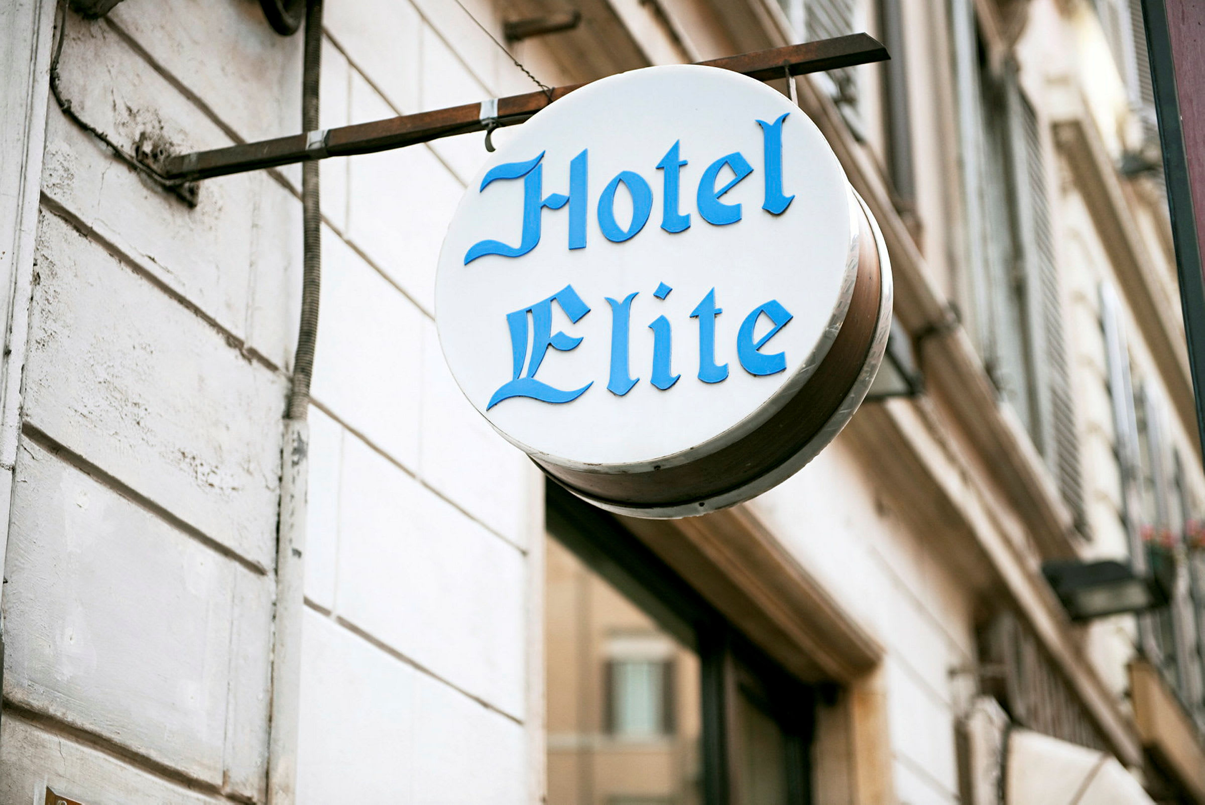 Et skilt med teksten "Hotel Elite" i blå bokstaver, montert på en hvit bygningvegg. (AI-generert tekst)