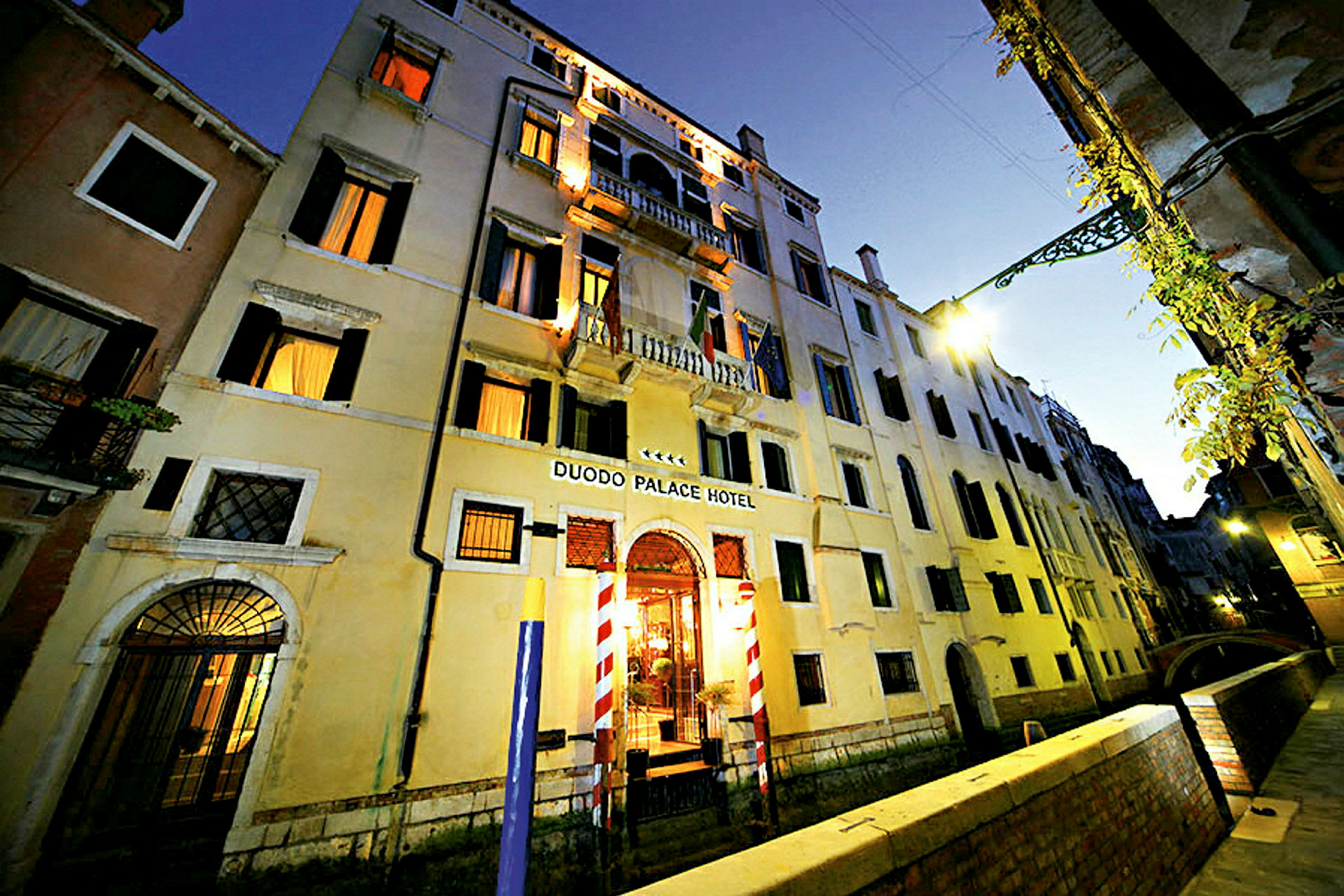 Leilighetsbygning i tre etasjer med skilt Duodo Palace Hotel, omgitt av en kanal og en gangvei. (AI-generert tekst)
