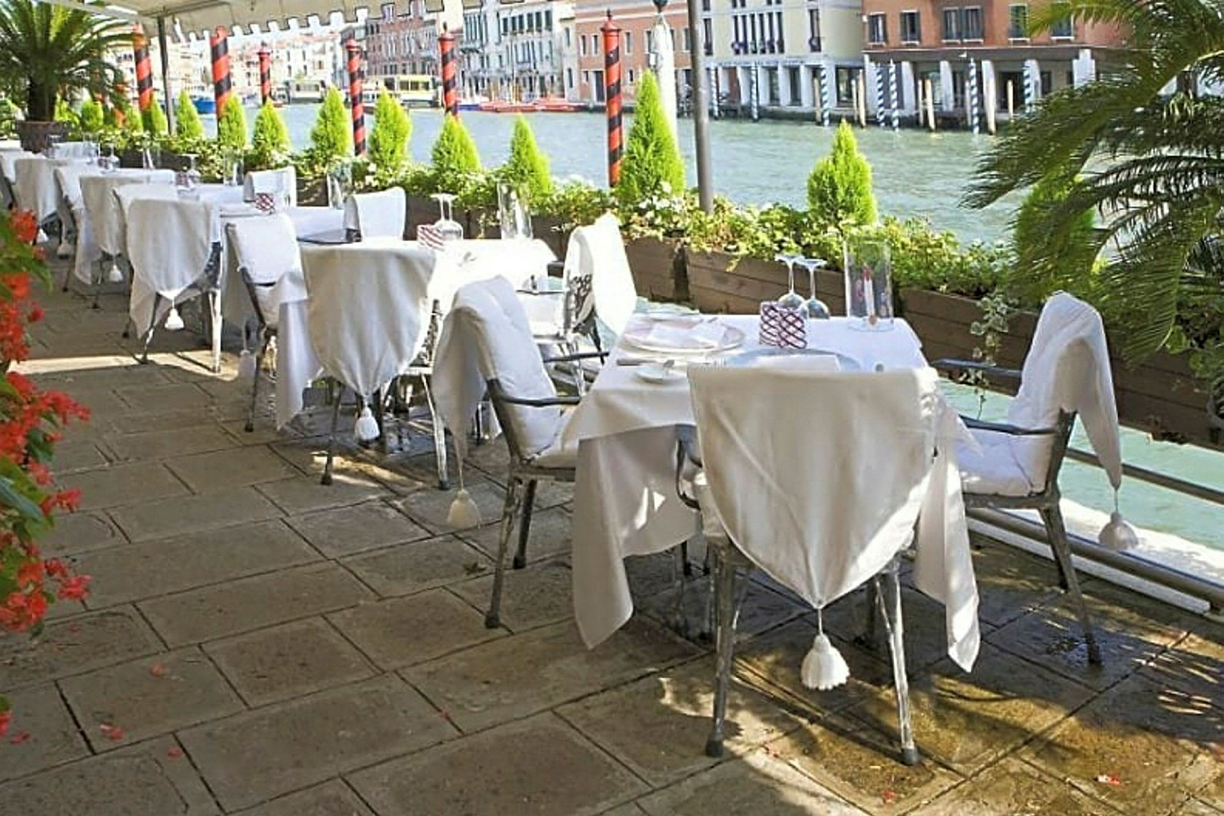 En uterestaurant med hvitdukte bord ved en kanal med flere bygninger i bakgrunnen. (AI-generert tekst)