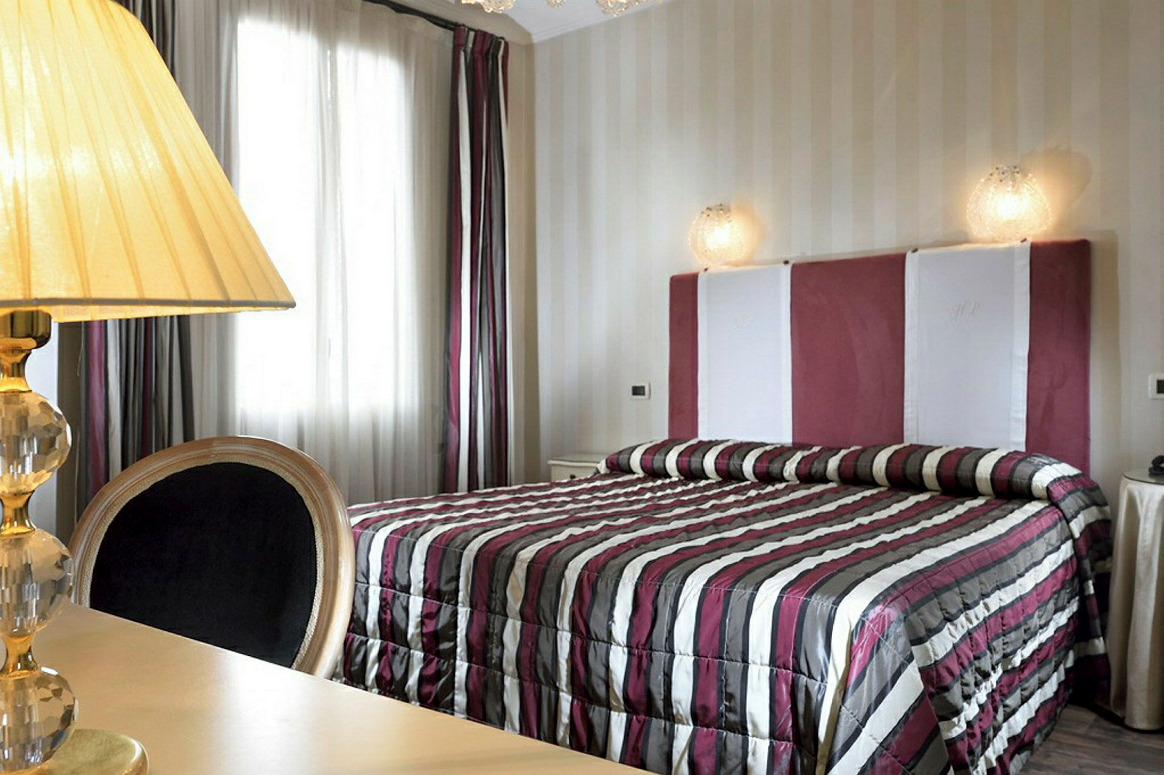 Et soverom med en seng som har et stripete sengetøy og en polstret sengegavl. Til venstre er det en lampe på et bord. (AI-generert tekst)