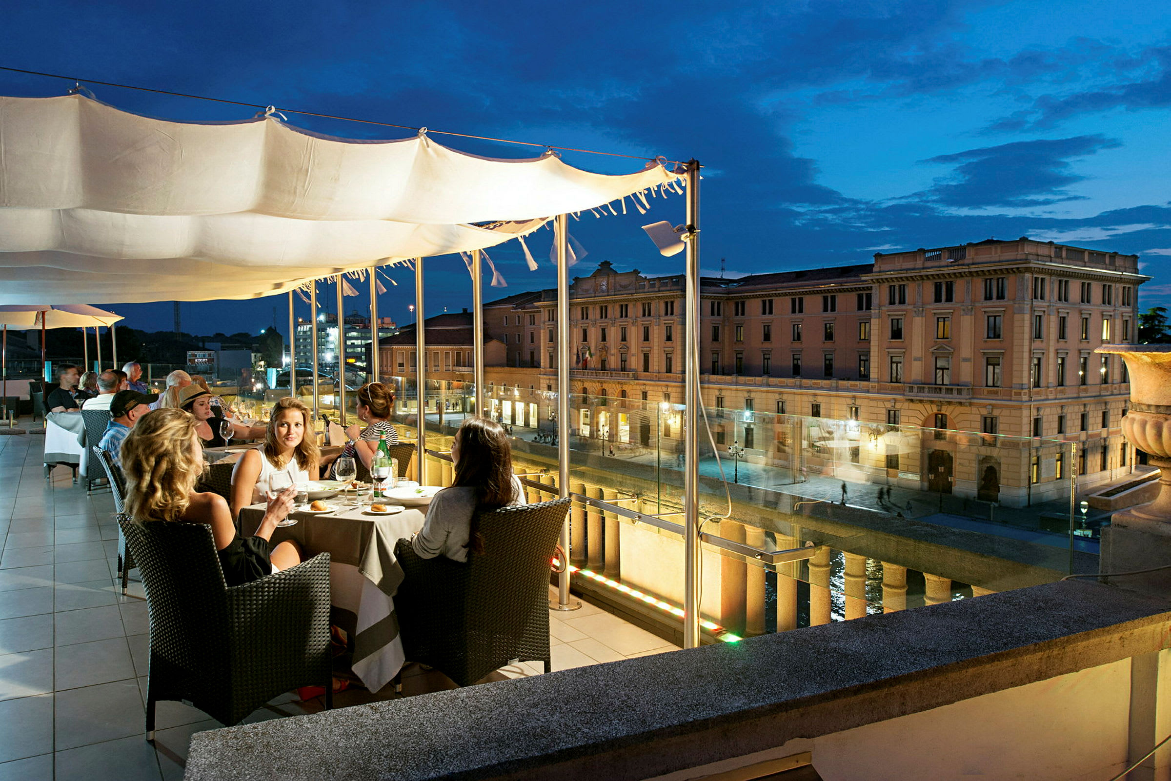 En restaurantterrasse om kvelden med utsikt over en bygate og en klassisk bygning i bakgrunnen. (AI-generert tekst)