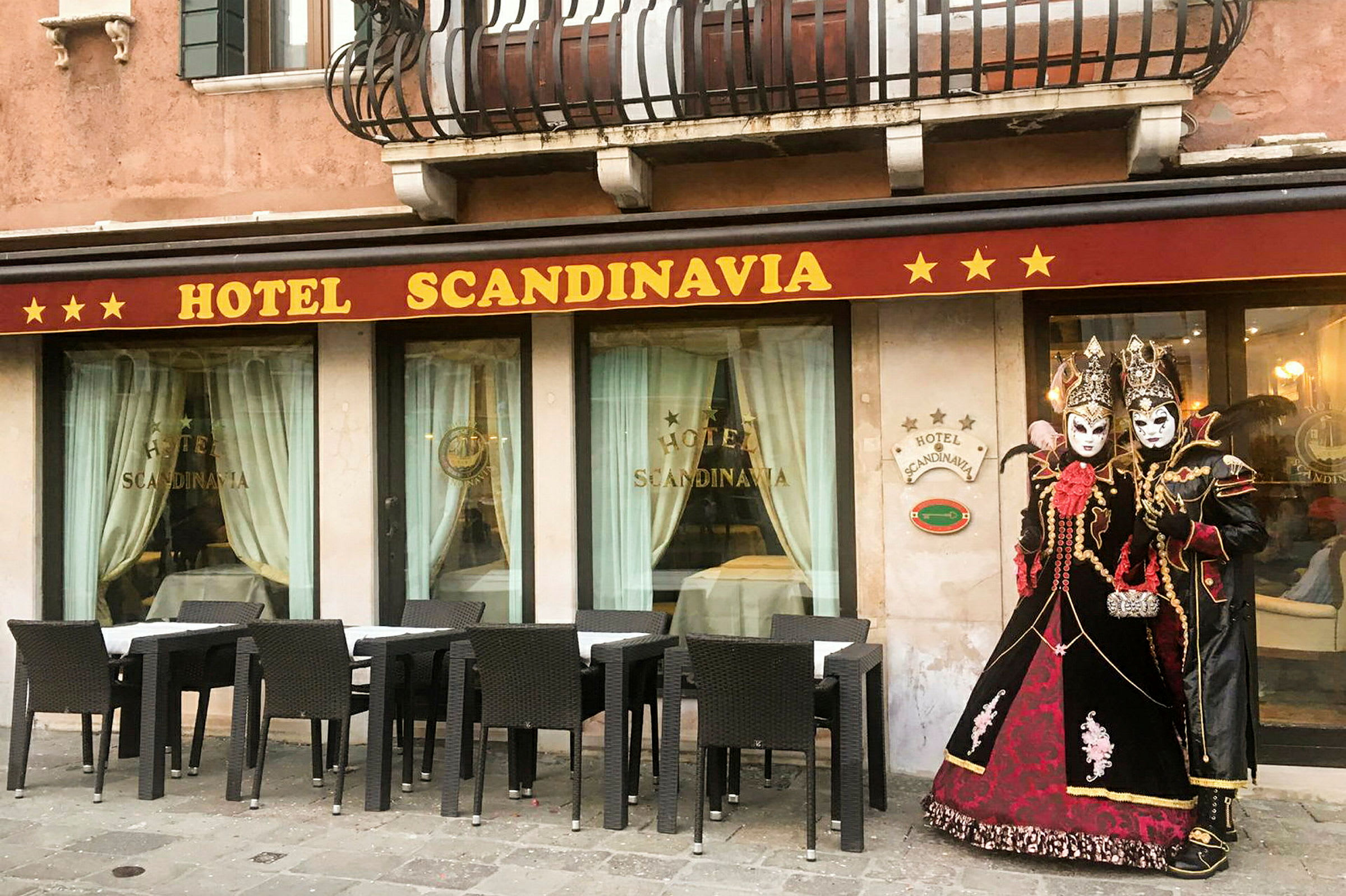 Hotellfasade med navnet 'Hotel Scandinavia', møblerte sitteplasser utendørs og personer kledd i tradisjonelle maskeradeklær. (AI-generert tekst)