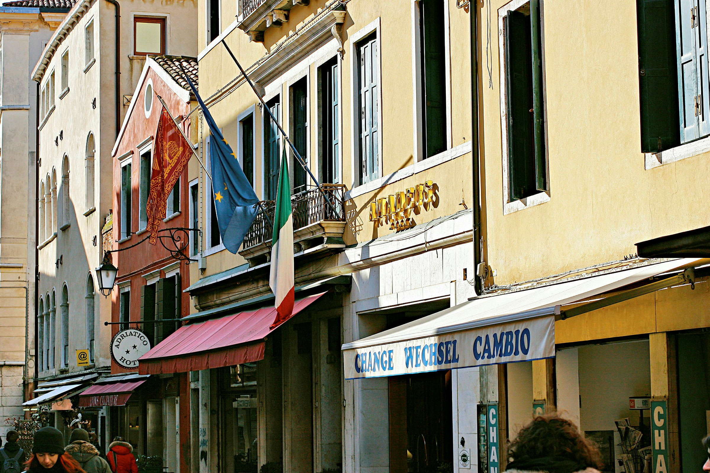 Fasade av bygninger med ett italiensk flagg, europeisk flagg og skilt som angir hotell og veksling. (AI-generert tekst)