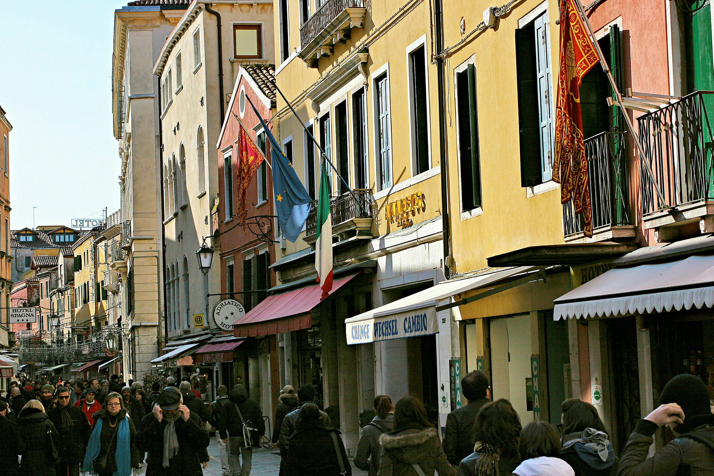 En travel gate omgitt av fargerike bygninger med italienske og europeiske flagg, omkranset av mennesker som går. (AI-generert tekst)