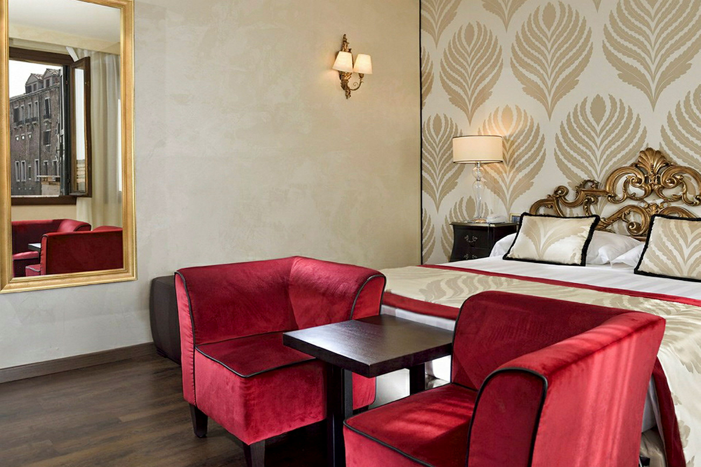 Et elegant hotellrom med en dobbeltseng, røde lenestoler og et lite bord, samt et vindu med utsikt. (AI-generert tekst)