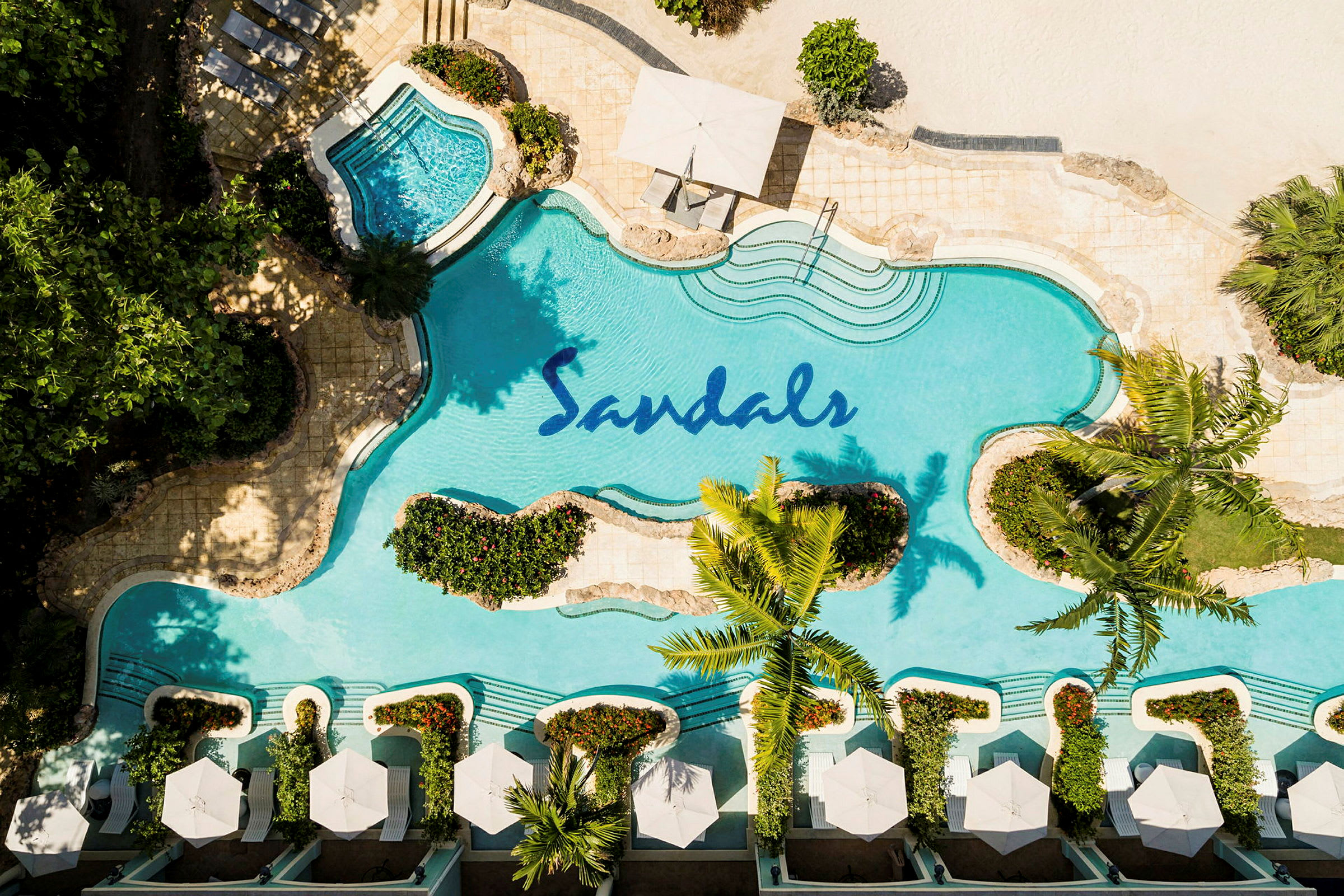 Et fugleperspektiv av et basseng med Sandals-logoen, omgitt av planter og solsenger med parasoller. (AI-generert tekst)