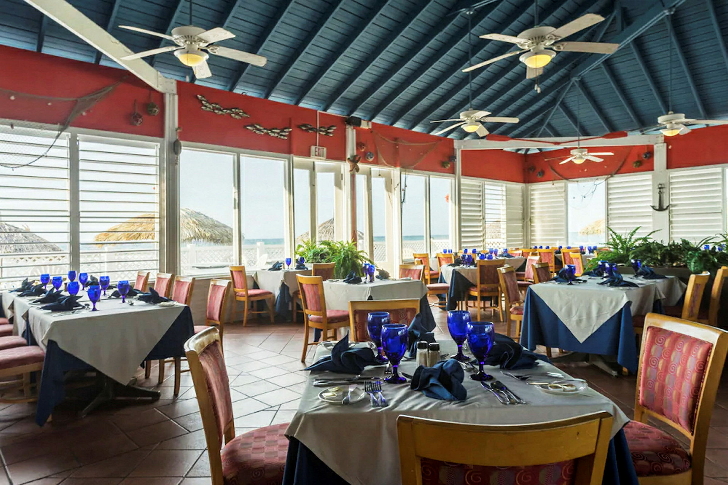 En restaurant med dekkede bord, blå glass og servietter, plassert i et rom med store vinduer og utsikt over havet. (AI-generert tekst)