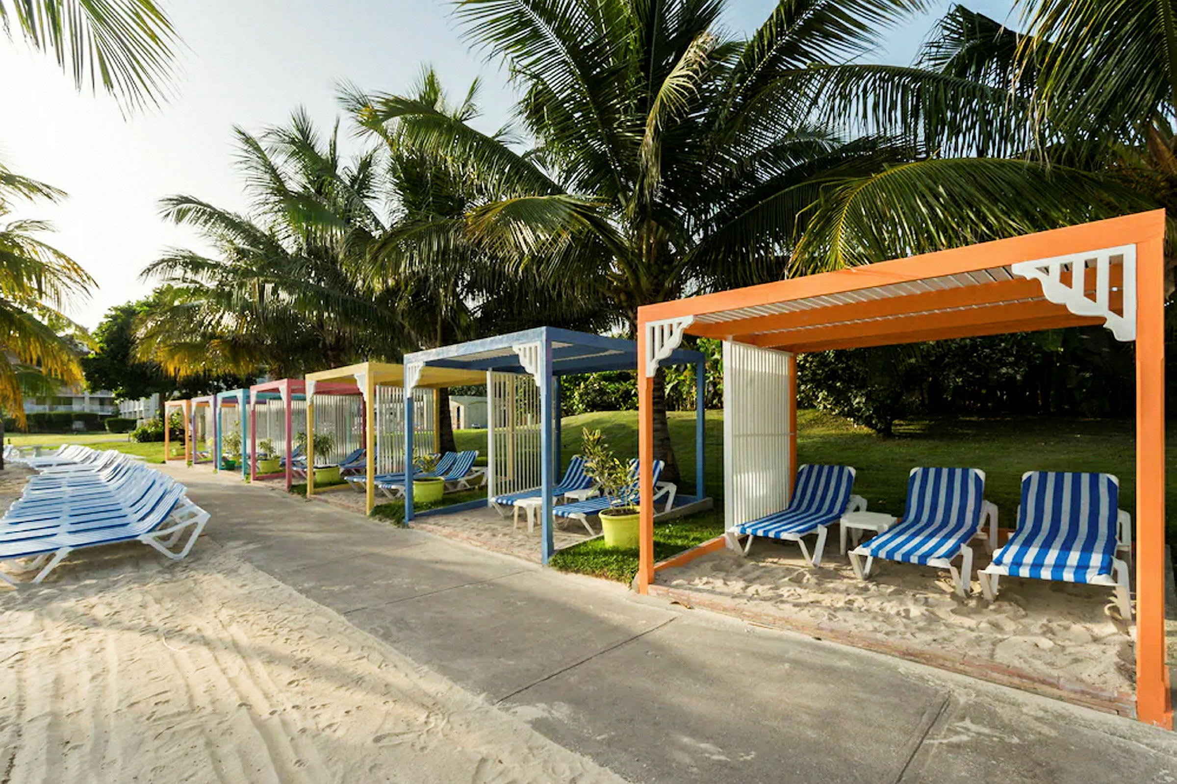 Flere fargerike paviljonger med solsenger under palmer ved en strandpromenade. (AI-generert tekst)