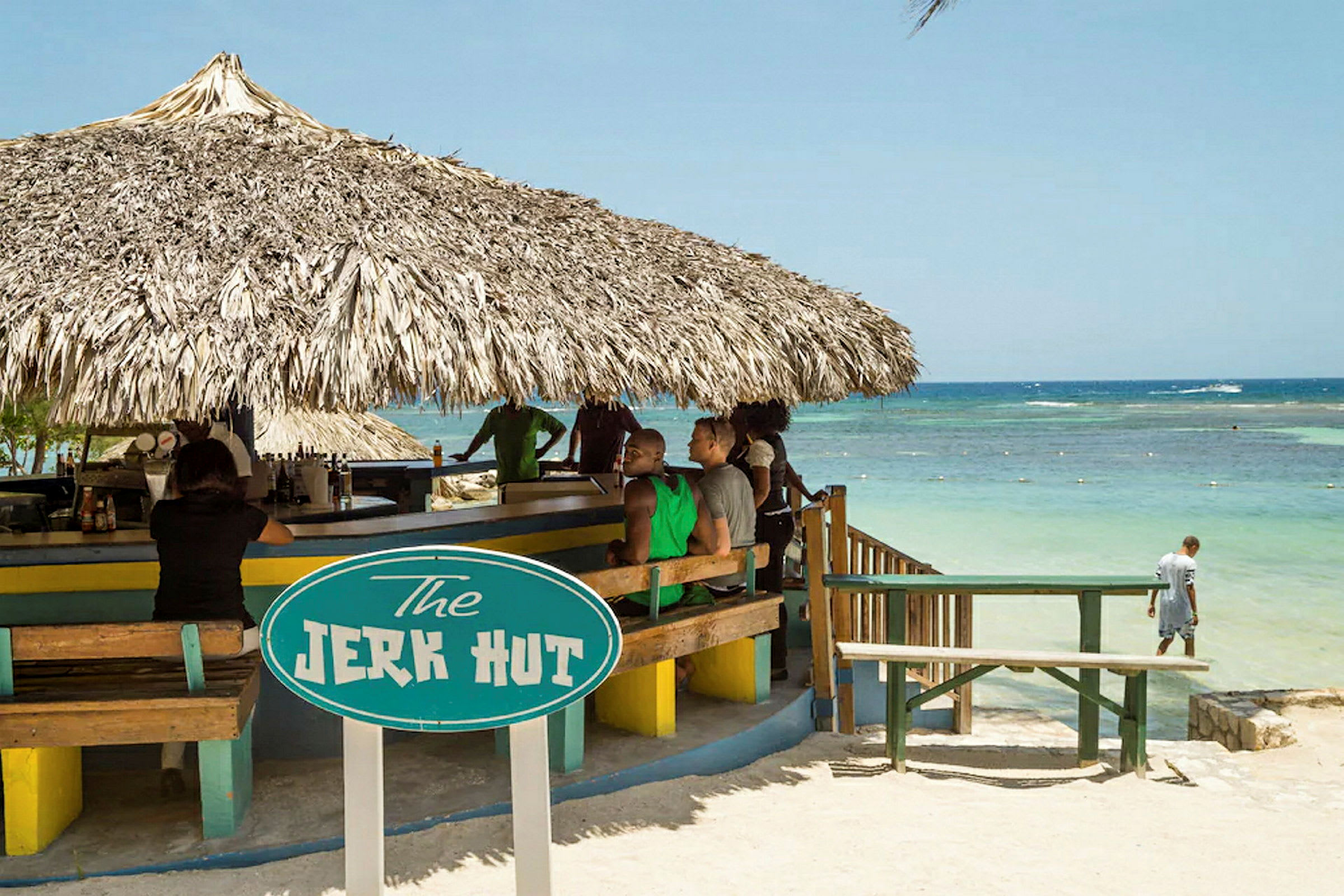 En strandbar med stråtak og et skilt som sier "The Jerk Hut". Havet er synlig i bakgrunnen. (AI-generert tekst)