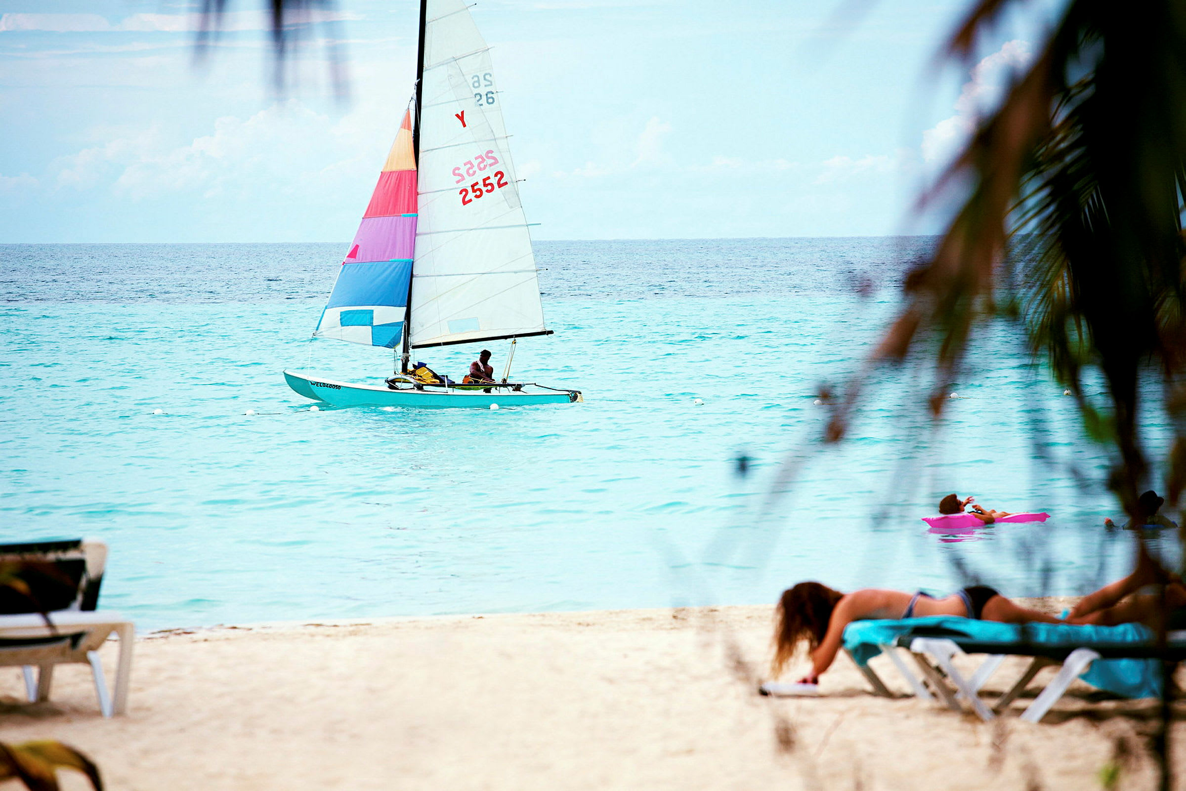 En seilbåt med fargerike seil på havet, med en person som ligger på en solseng på en sandstrand i forgrunnen. (AI-generert tekst)
