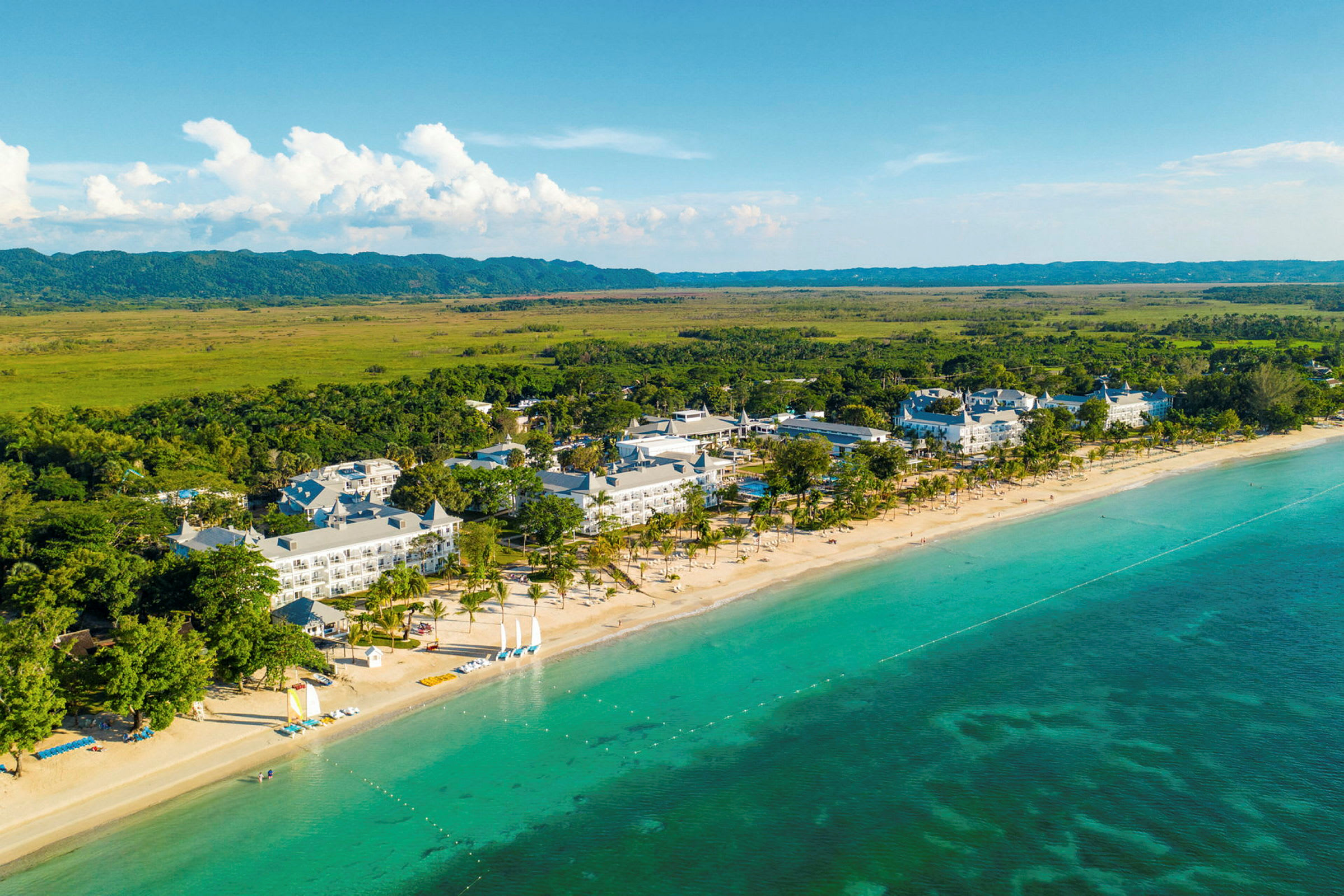 Flyfoto av et kystnært hotellområde med sandstrand, klart turkis vann og omkringliggende vegetasjon. (AI-generert tekst)