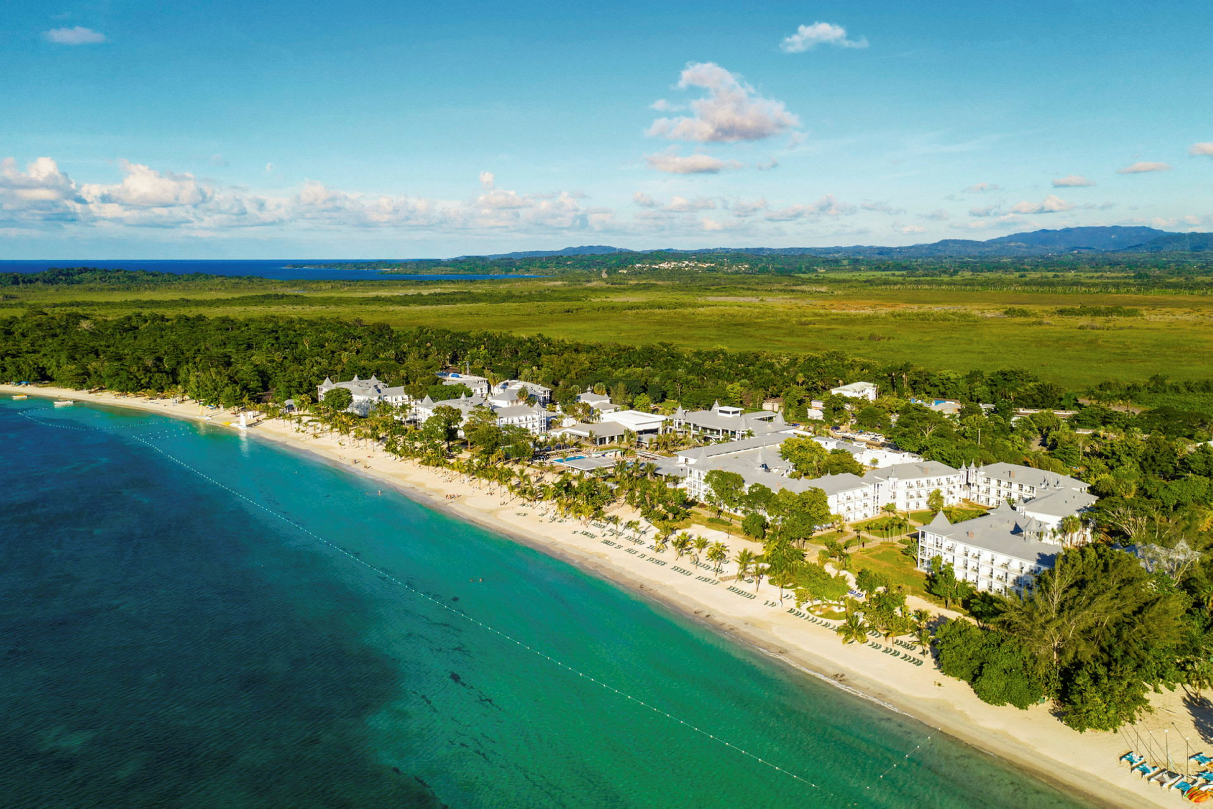 Luftfoto av en strand ved et turkist hav og et hotellkompleks omgitt av frodig grøntområde. (AI-generert tekst)