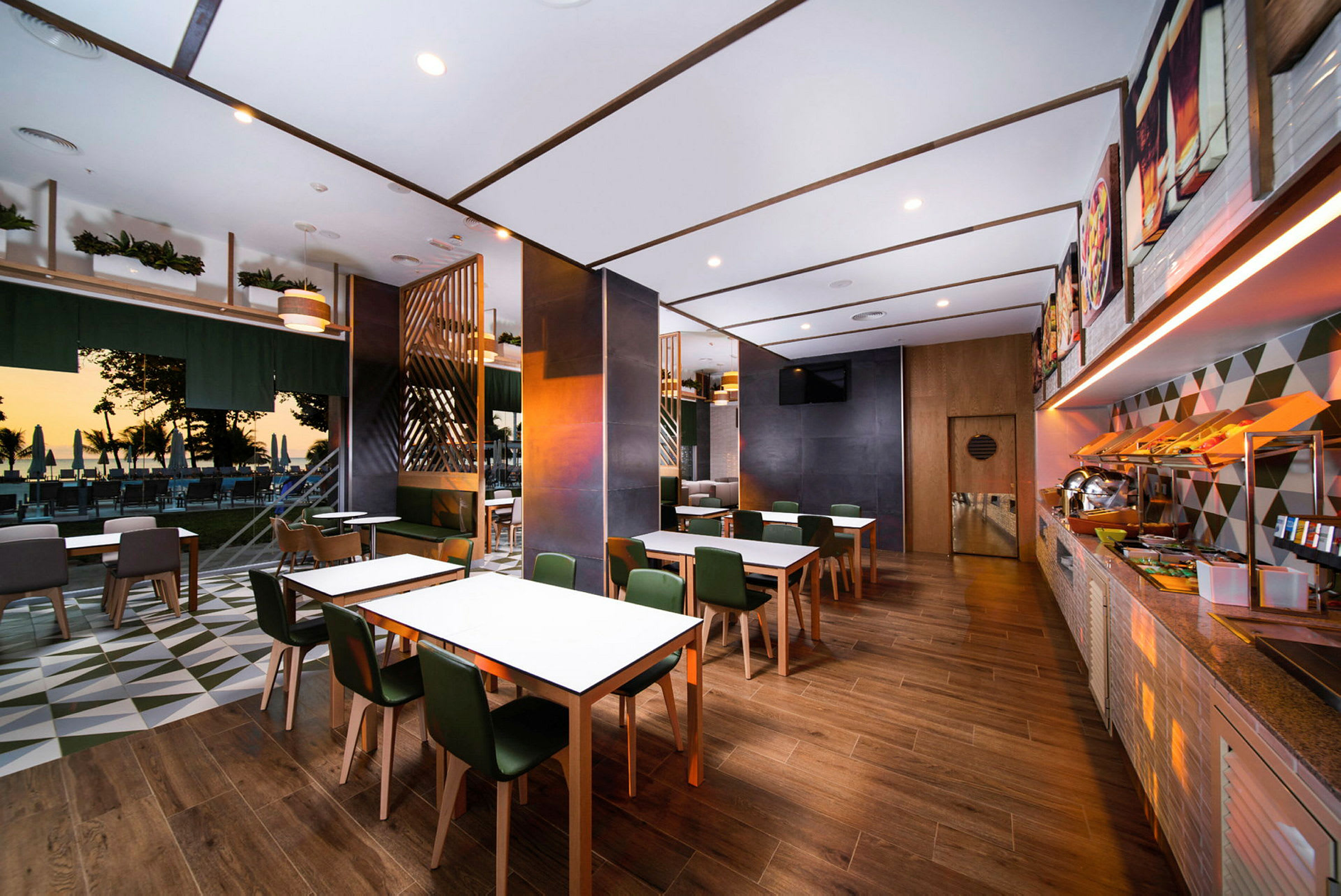 Et moderne restaurantområde med tremøbler, hvite bord og grønne stoler. I bakgrunnen er en utendørs serveringsplass med utsikt mot stranden ved solnedgang. (AI-generert tekst)