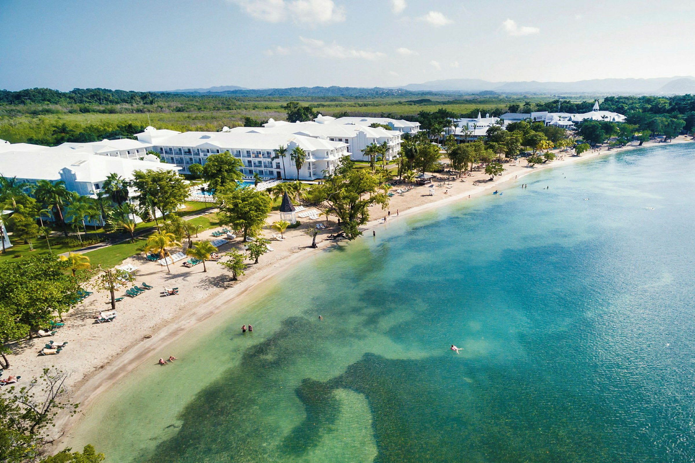 Bilde av en strand med klart blått vann og hotellbygninger i bakgrunnen. (AI-generert tekst)
