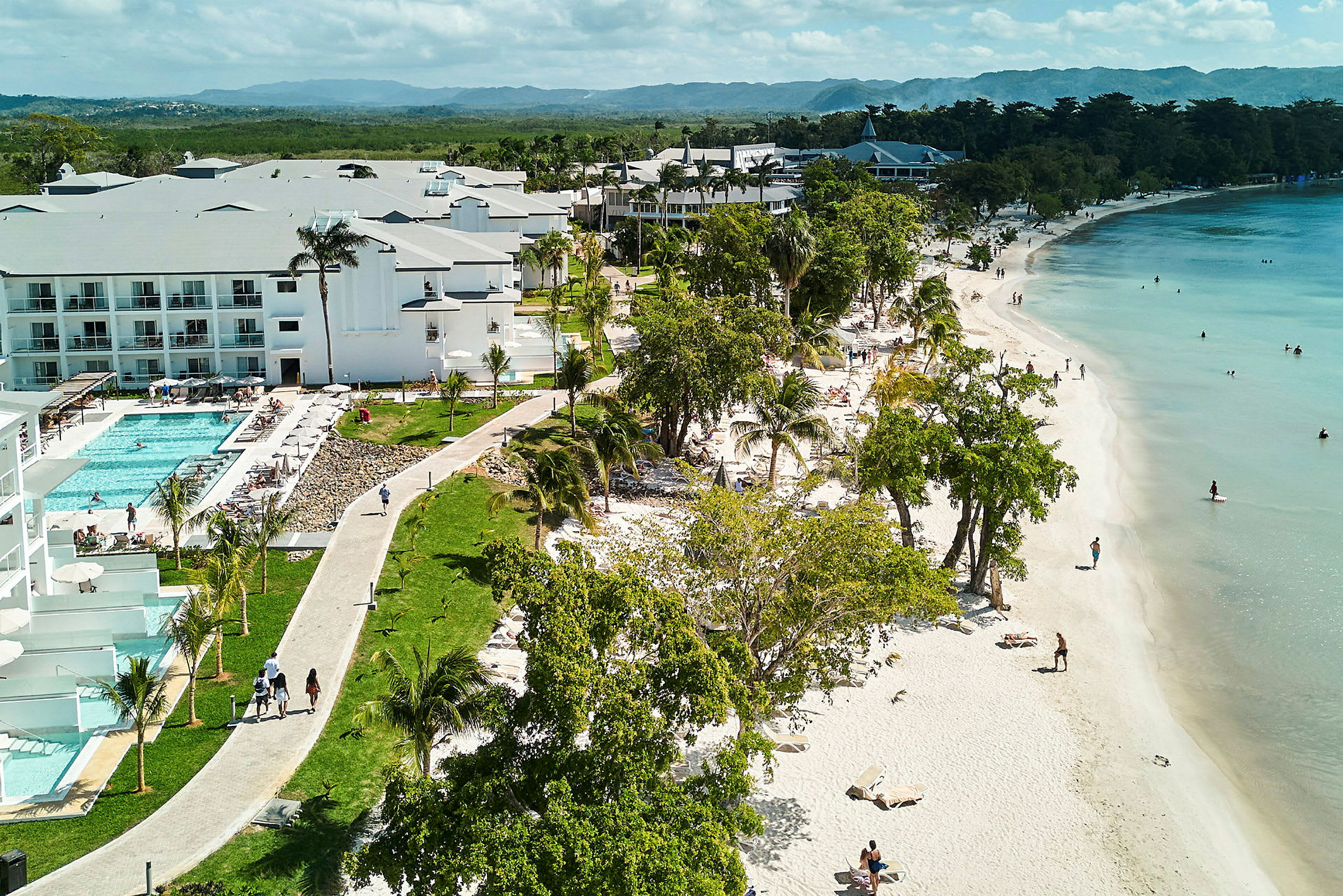En sandstrand med personer som svømmer i sjøen og hotellbygninger ved siden av et basseng. (AI-generert tekst)