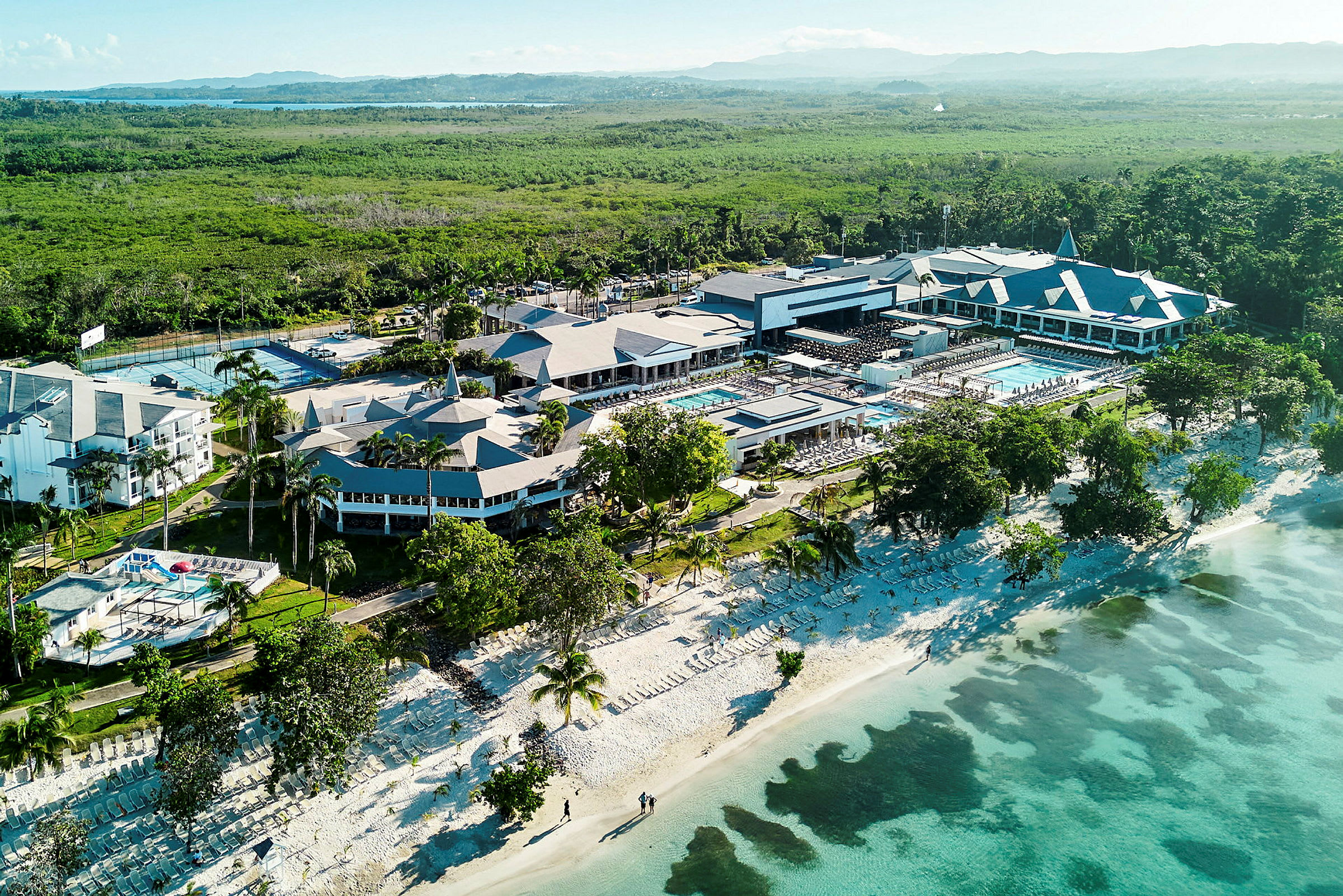 Luftfoto av en resort ved stranden med bassengområder og omkringliggende grøntområder. (AI-generert tekst)