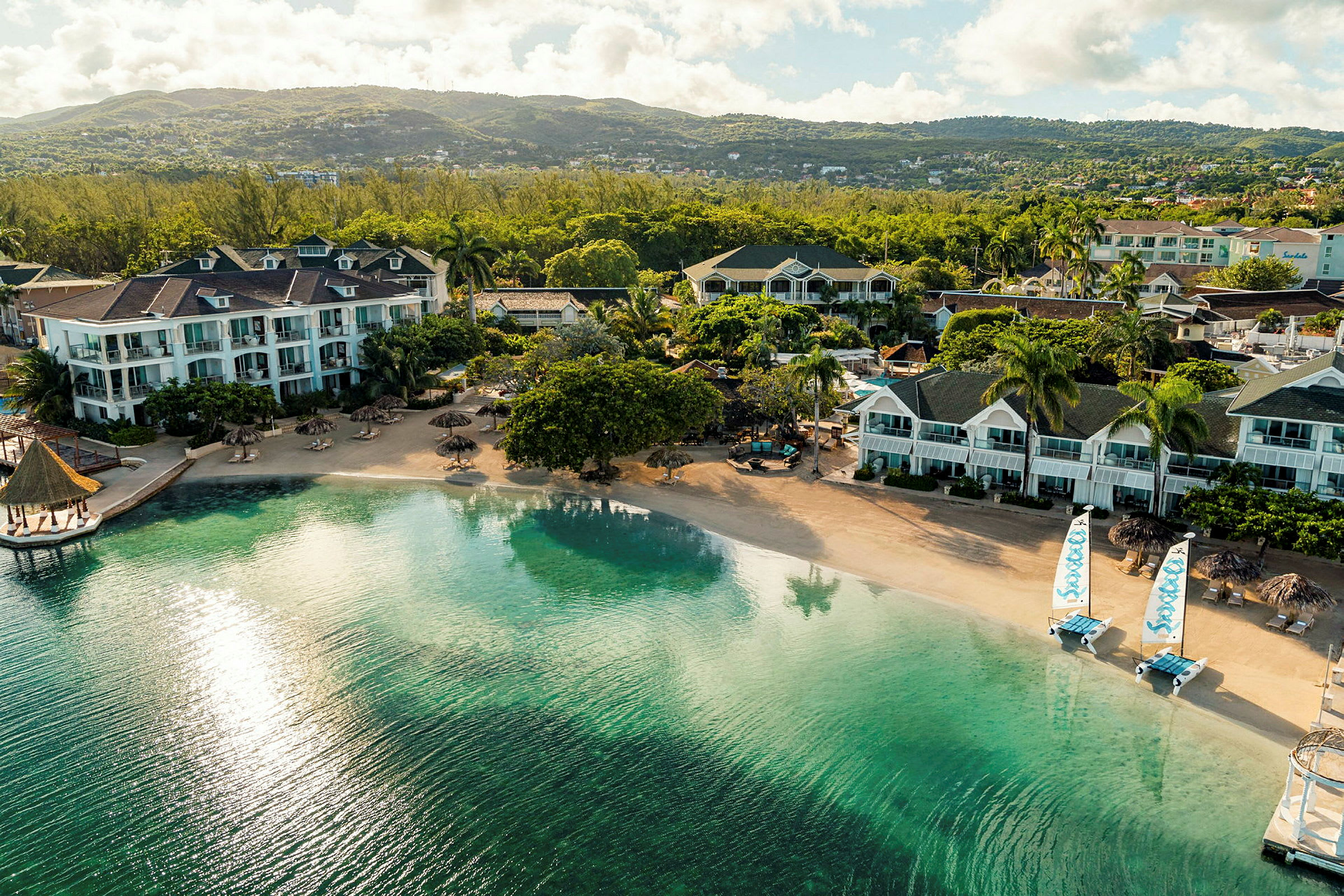 Flere hotellbygninger ved en rolig sandstrand med klart vann og utsikt over frodige åser. (AI-generert tekst)