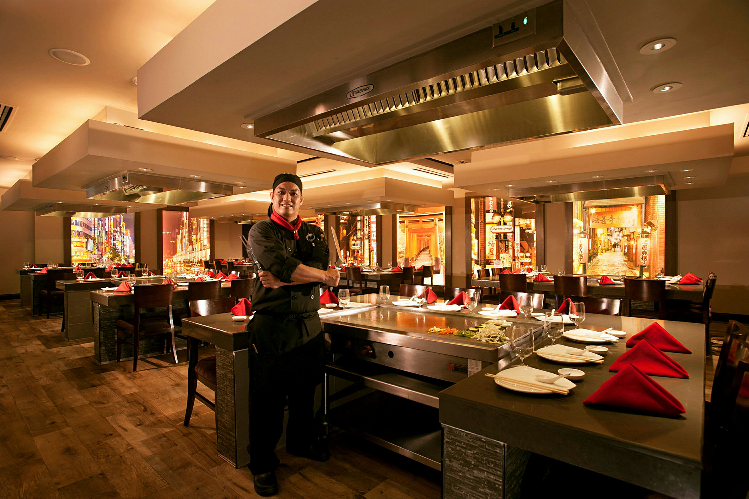 Interiør av en restaurant med oppstilte bord og stoler. Et teppanyaki-kjøkken er synlig med kjøkkenutstyr og ingredienser. (AI-generert tekst)