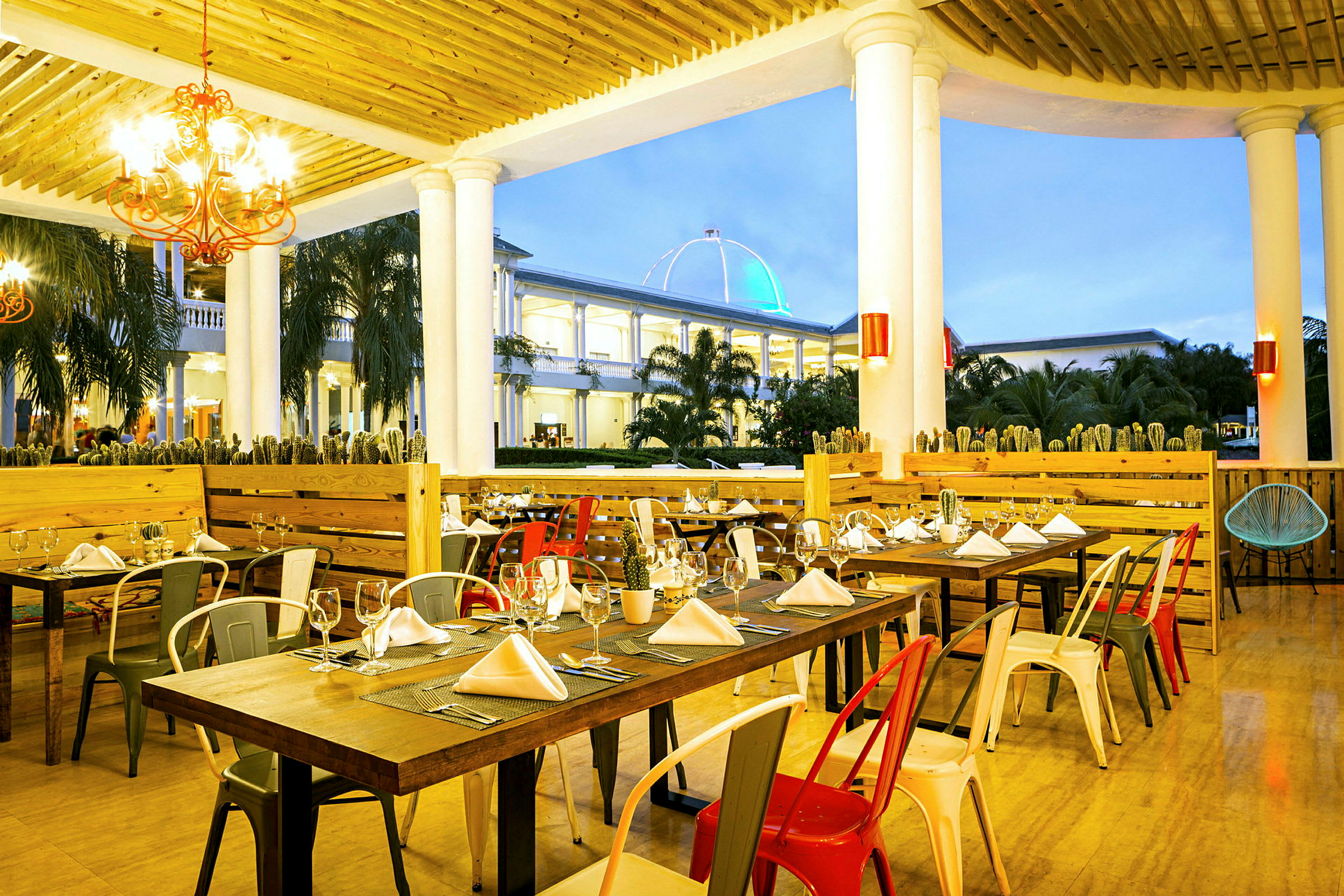 Bilde av en restaurant med dekkede bord og fargerike stoler, plassert under en trekonstruksjon med søyler. I bakgrunnen vises en bygning med et opplyst kuppeltak. (AI-generert tekst)
