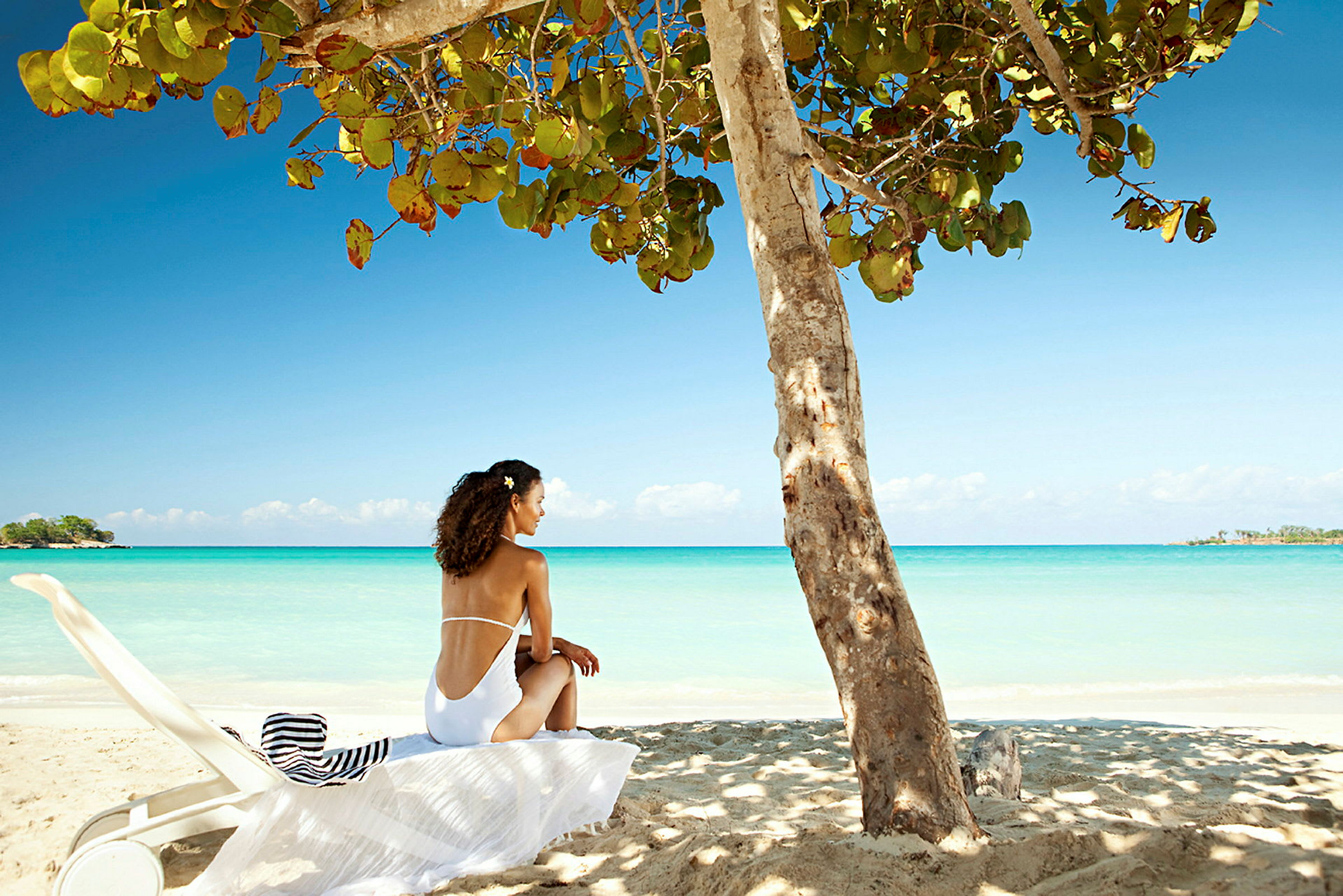 En person sitter på en solseng i skyggen av et tre ved en strand med klart blått hav i bakgrunnen. (AI-generert tekst)
