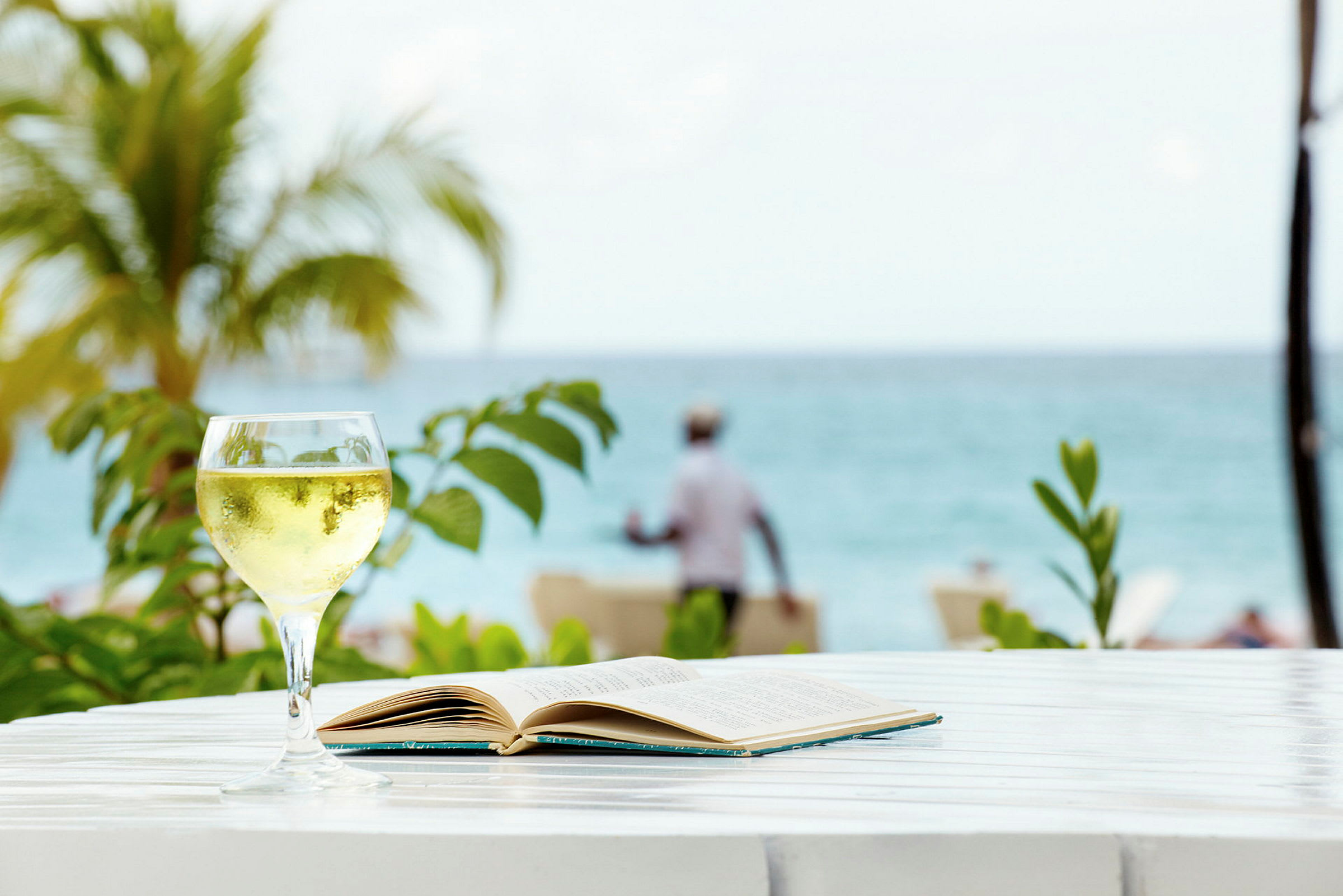 Et glass med drikke og en bok ligger på et bord med havet og et palme i bakgrunnen. (AI-generert tekst)