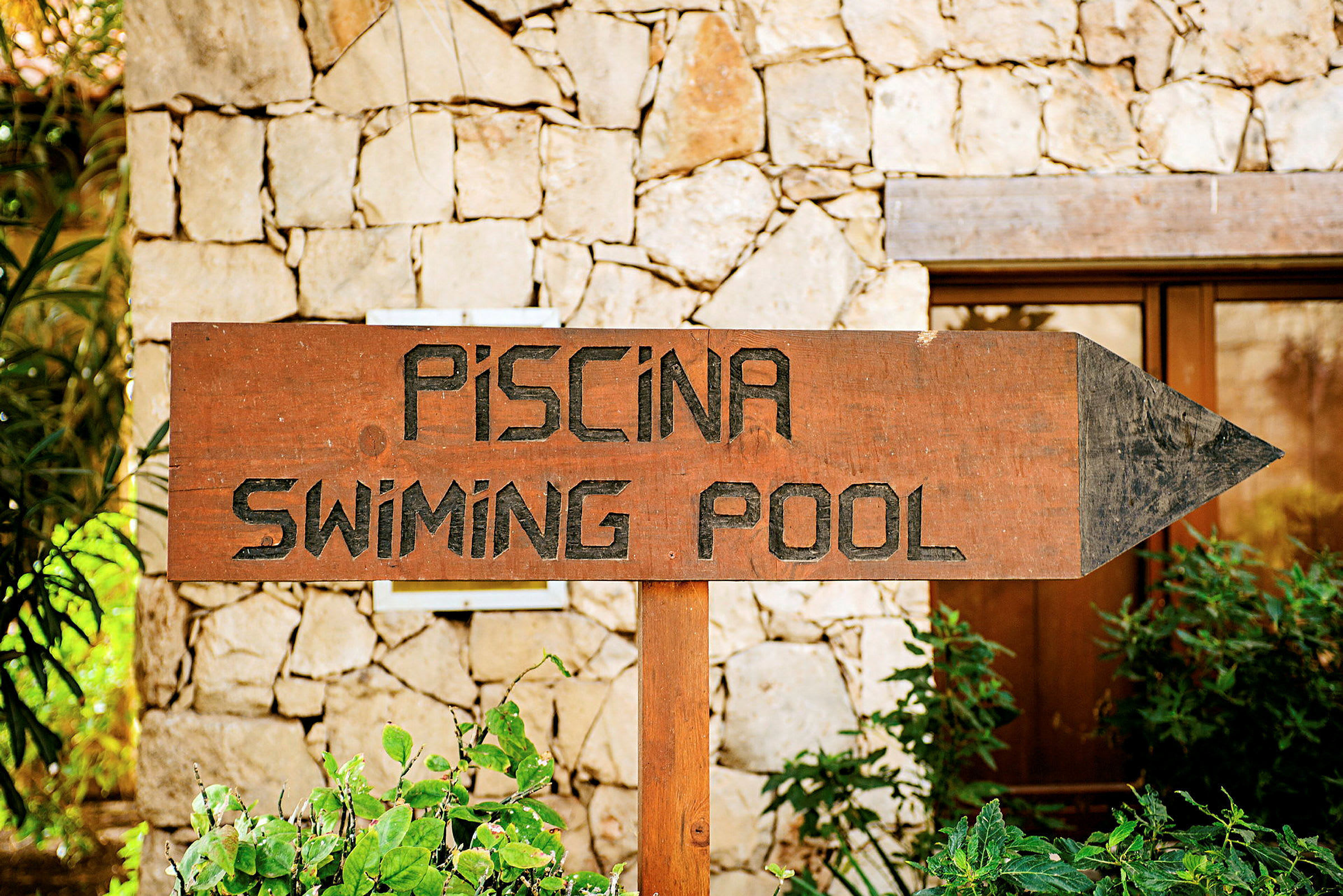 Skilt med teksten 'Piscina - Swimming Pool' foran steinmur. (AI-generert tekst)