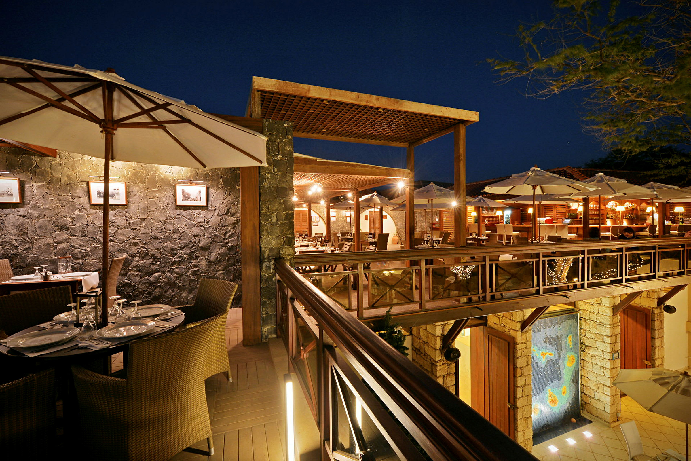 En godt opplyst restaurant med bord, stoler og parasoller på en treterrasse om kvelden. (AI-generert tekst)