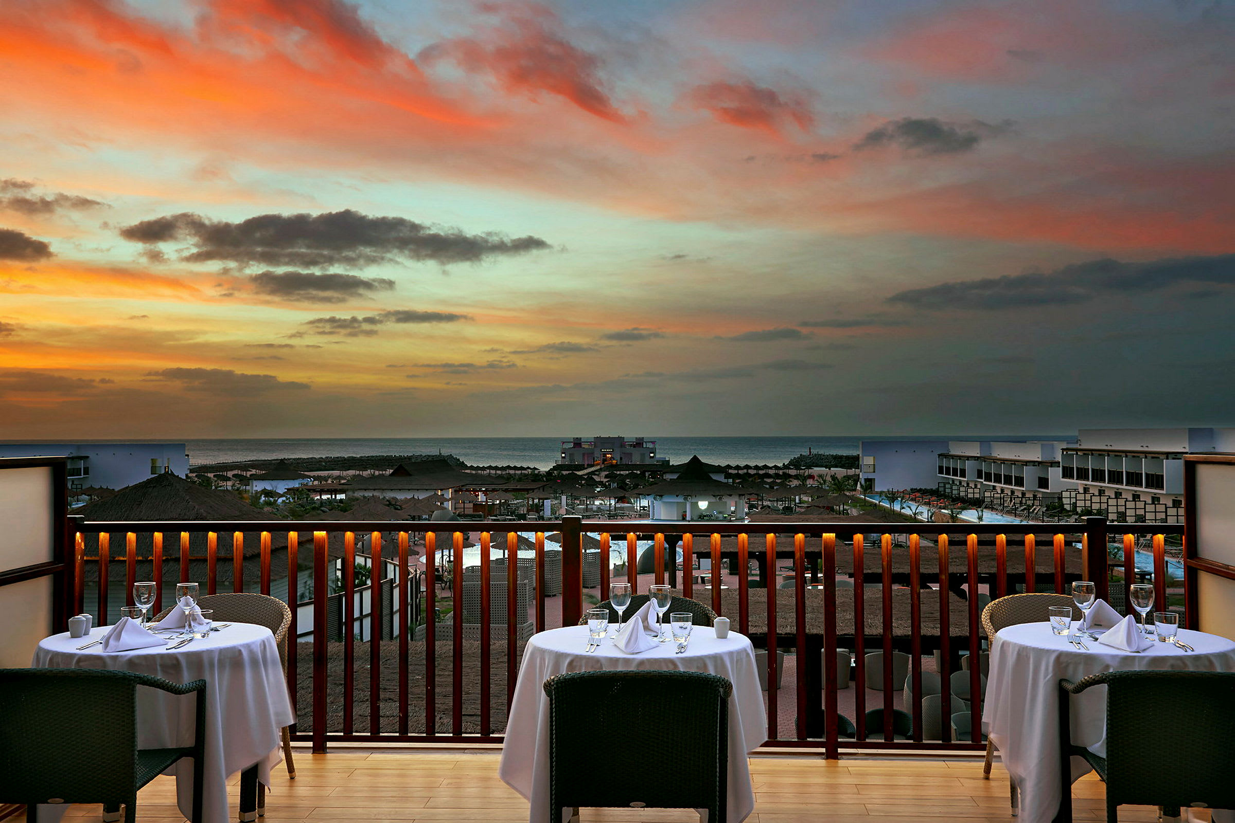 Utsikt fra en restaurant med dekkede bord mot solnedgangen over havet. (AI-generert tekst)