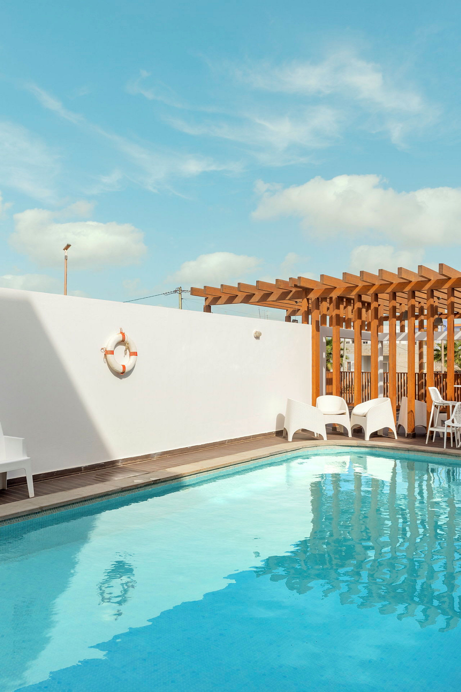 Utendørs svømmebasseng med hvit vegg og trepergola i bakgrunnen. (AI-generert tekst)