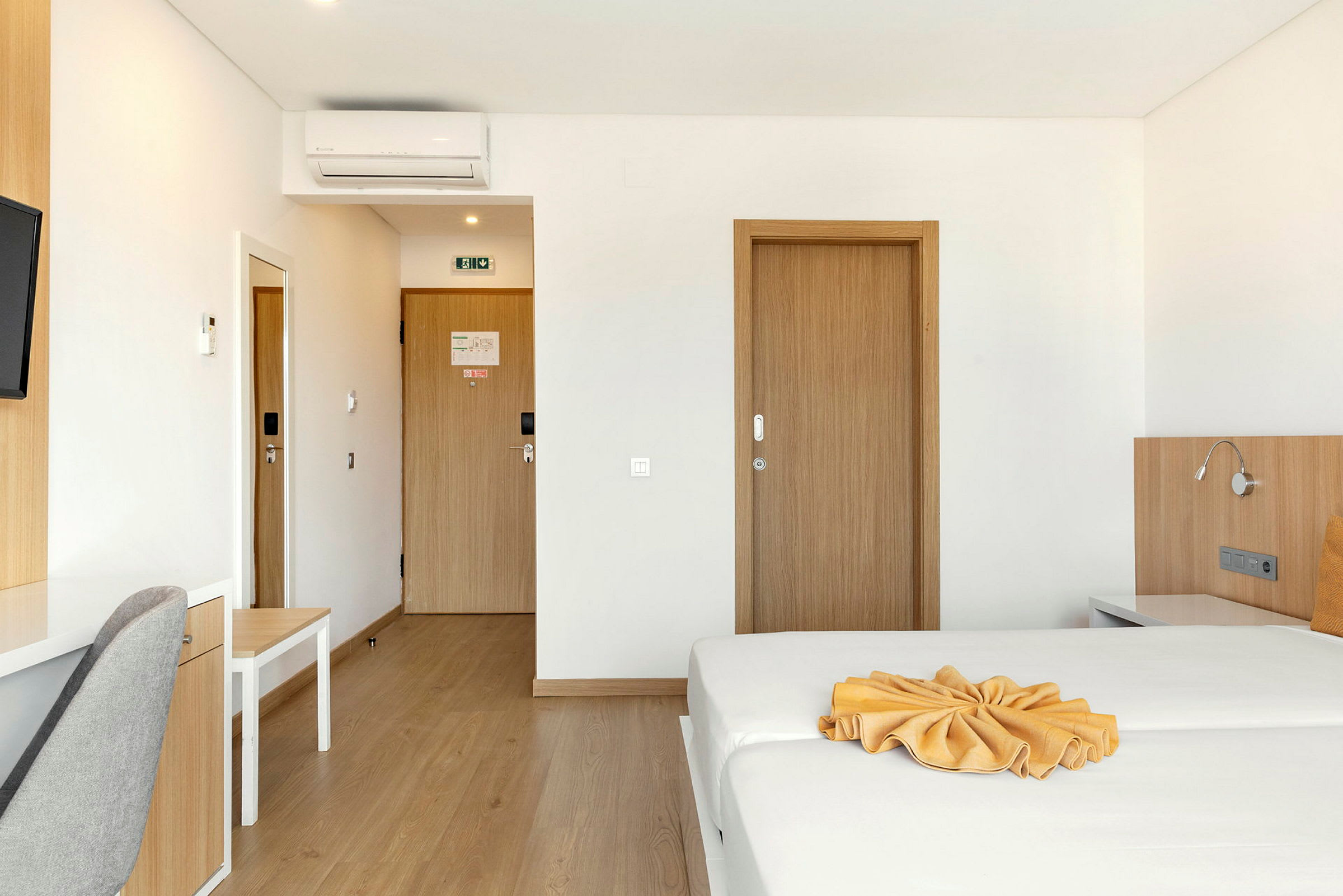 Et hotellrom med en oppredd seng og et skrivebord ved siden av en grå stol, tredører og en luftkondisjonering på veggen. (AI-generert tekst)