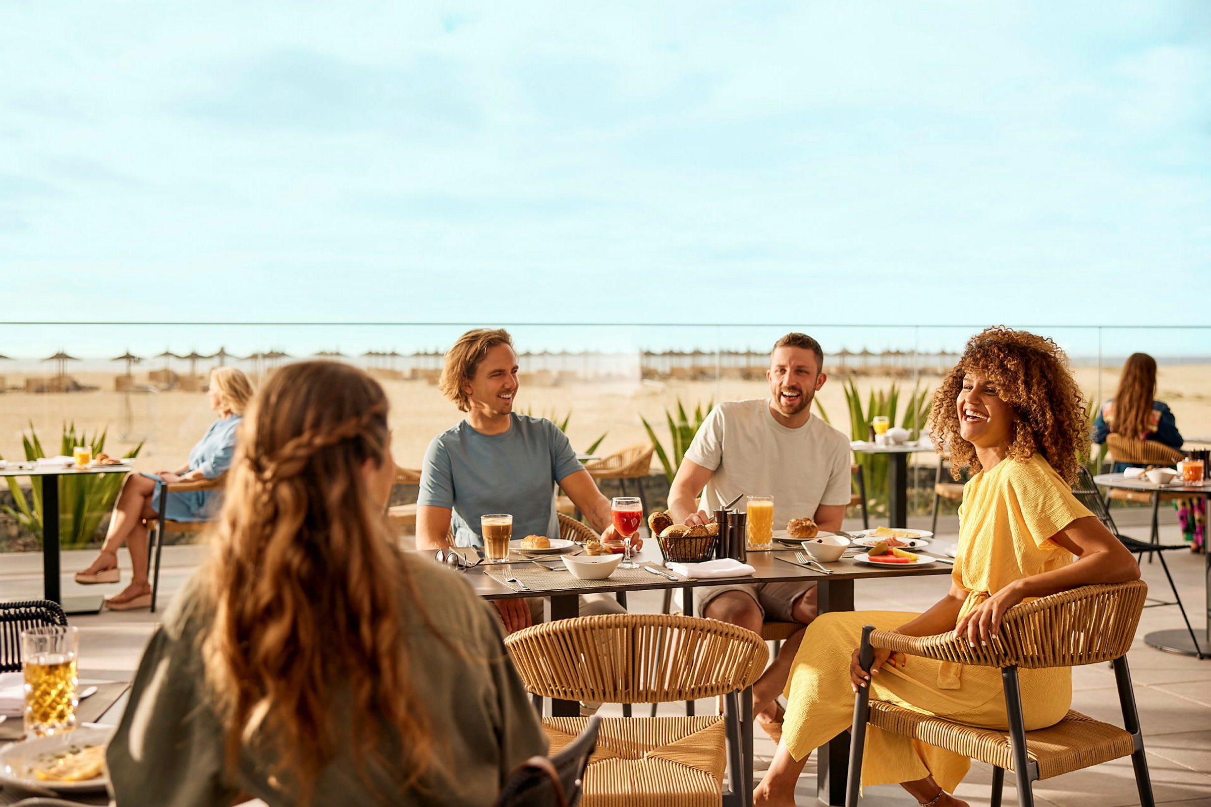 Folk sitter ved bord ute og spiser måltider, med en strand og havet i bakgrunnen. (AI-generert tekst)